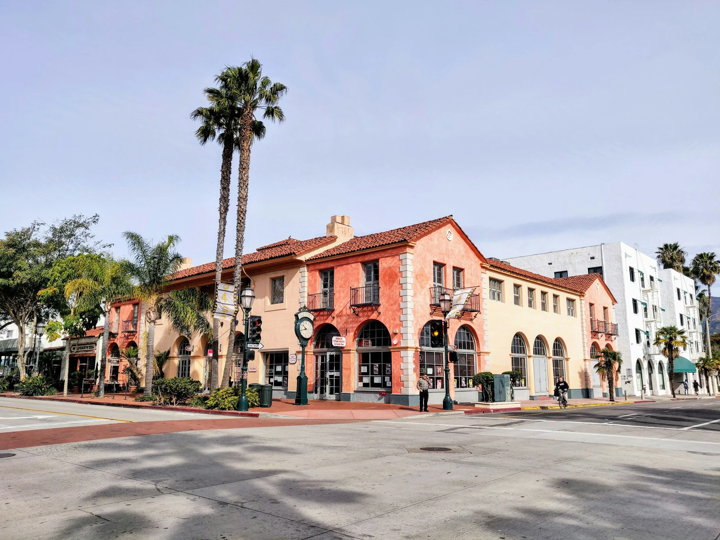 A street corner with a pink and beige European-style building featuring arched windows and balconies, palm trees, and a clear blue sky.