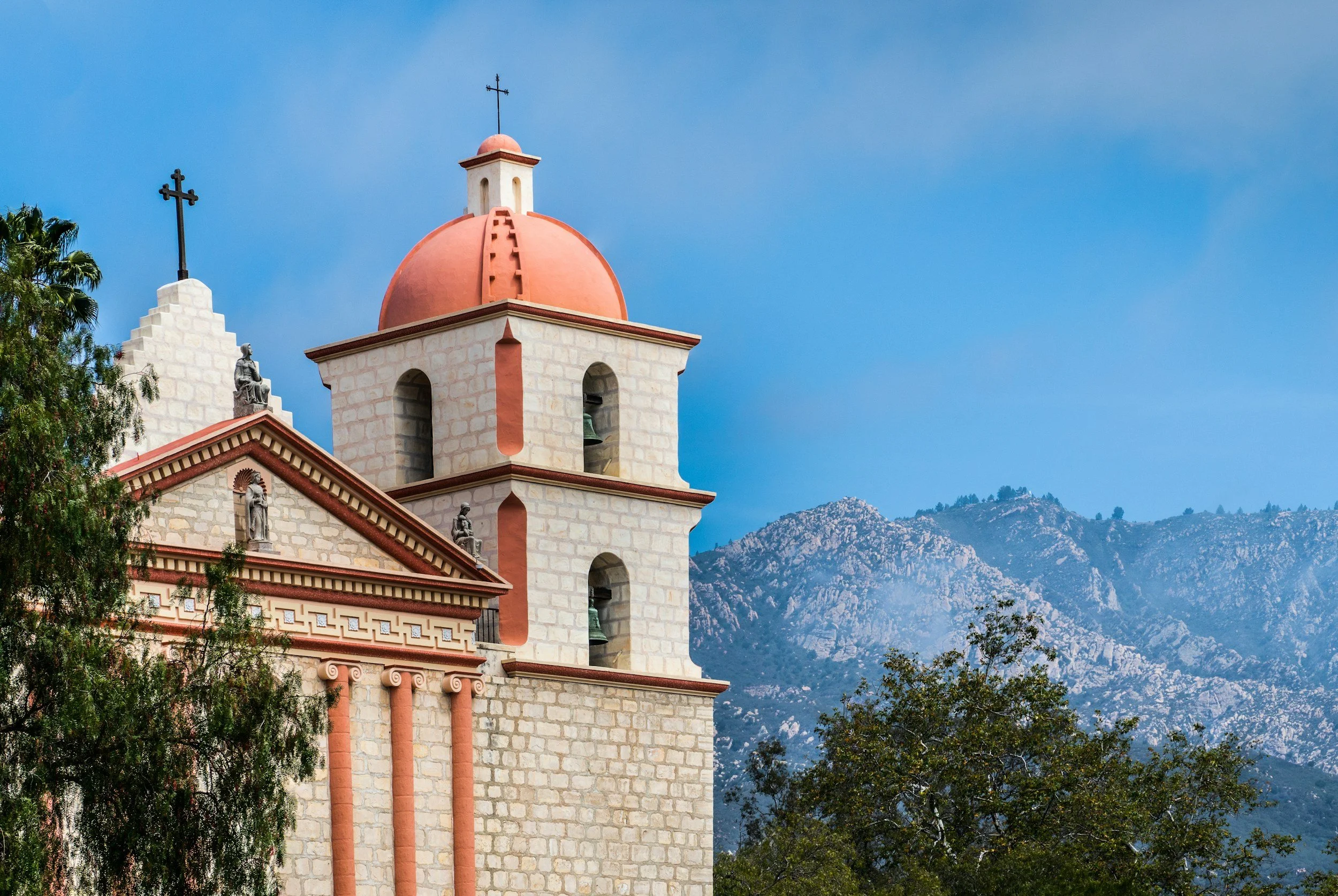 The image shows the top part of a stone church with a red dome and multiple statues along the facade, set against a background of mountains and a clear blue sky, with some trees visible at the bottom.