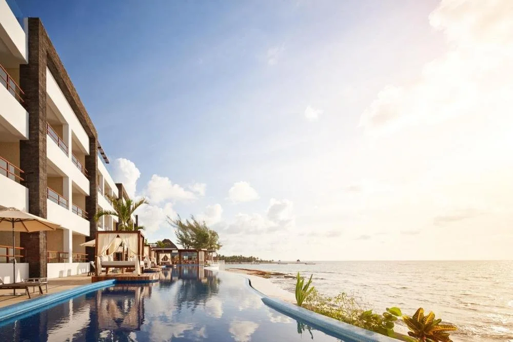 Beach resort with an infinity pool overlooking the ocean, hotel balconies, lounge chairs, umbrellas, and tropical plants, under a partly cloudy sky.