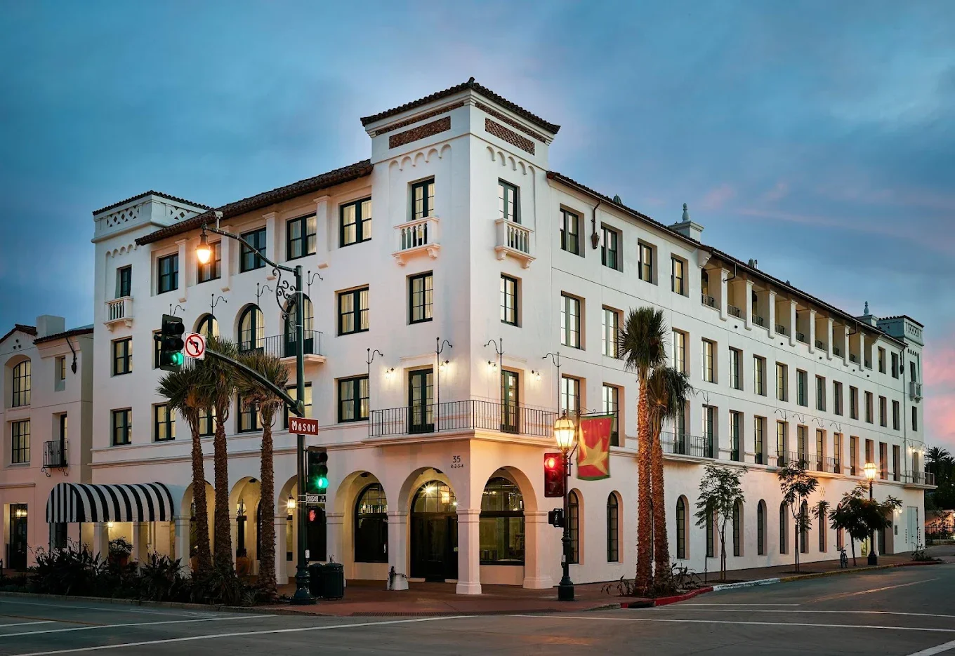 A white multi-story building with arched windows and balconies, located at a street corner, with palm trees and street lights, under a twilight sky.