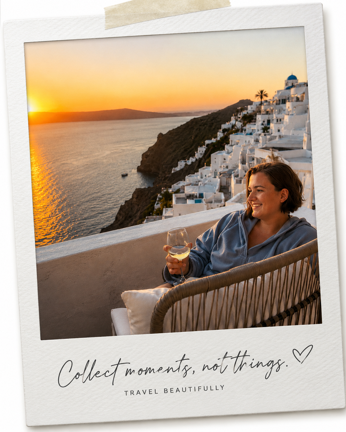 A woman sitting on a balcony holding a glass of white wine, overlooking a sunset over the ocean with white buildings and a hillside in Santorini, Greece.