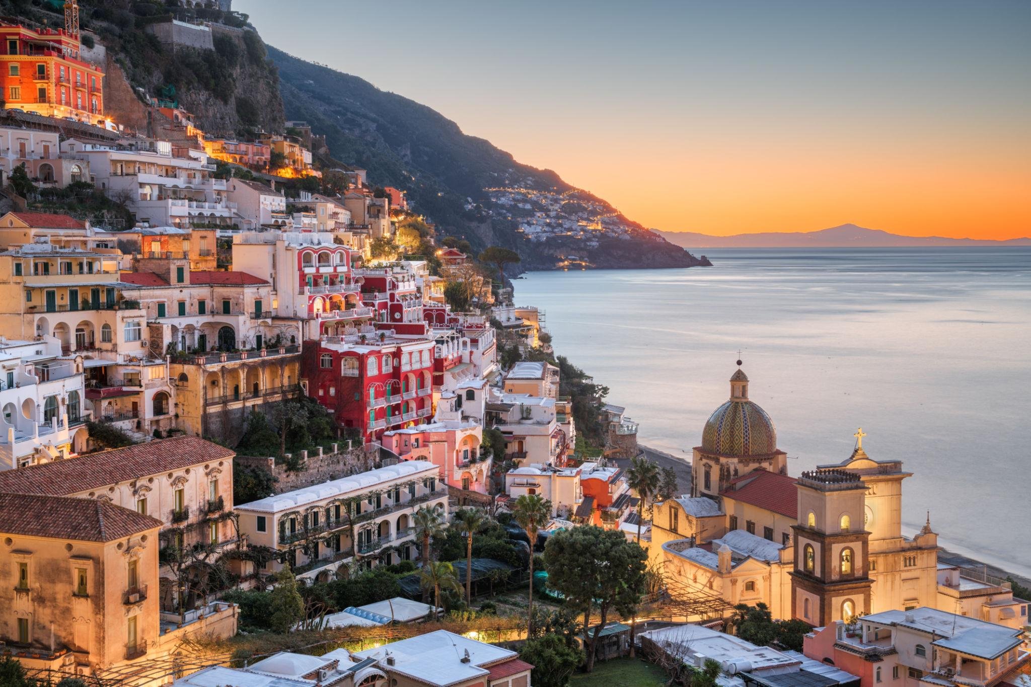 Colorful houses and buildings climbing hillside at sunset, overlooking the sea with a distant mountain in view.