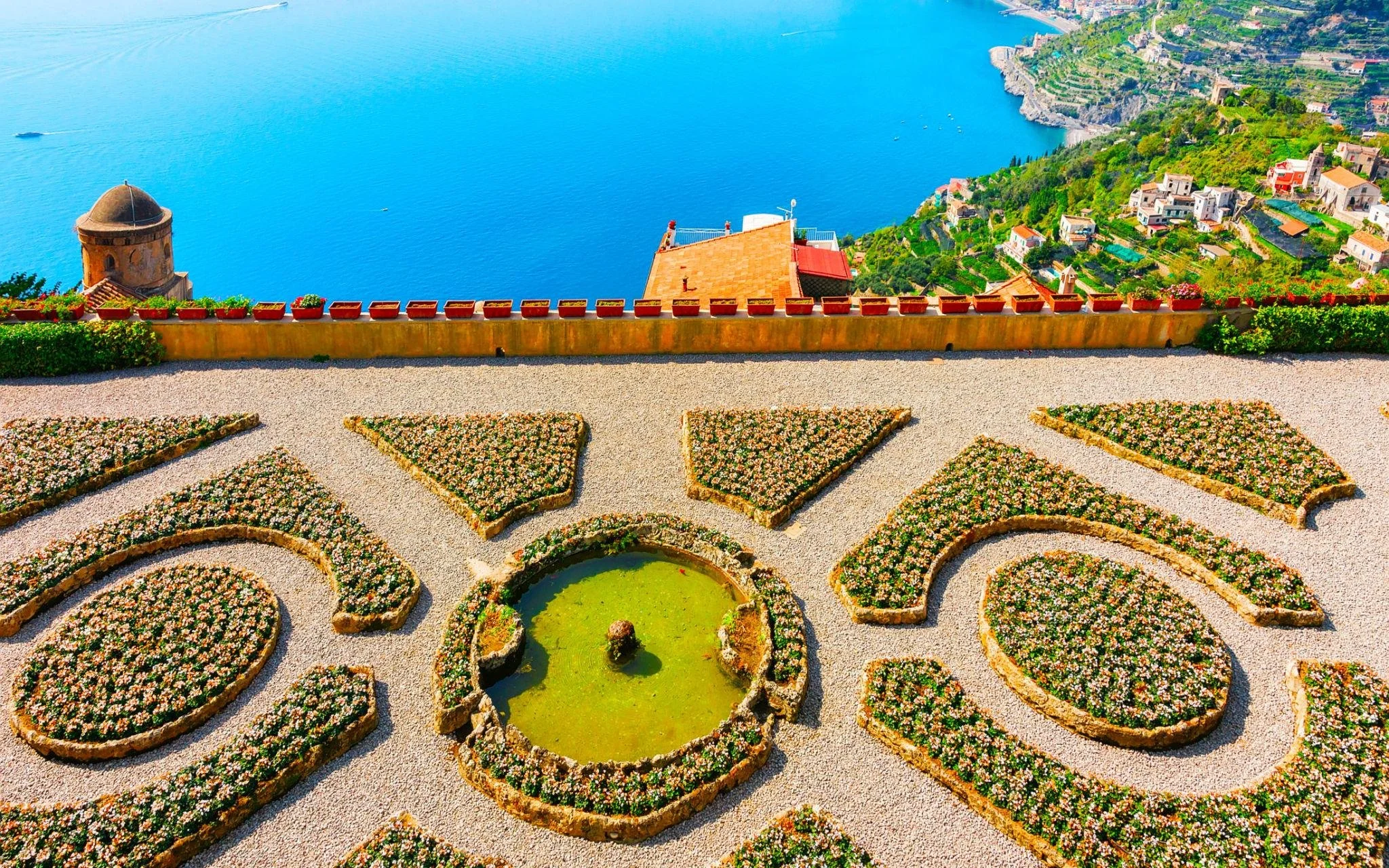 A terraced garden with symmetrical flower beds and a circular pond filled with green water, overlooking a large blue lake and a hillside village.
