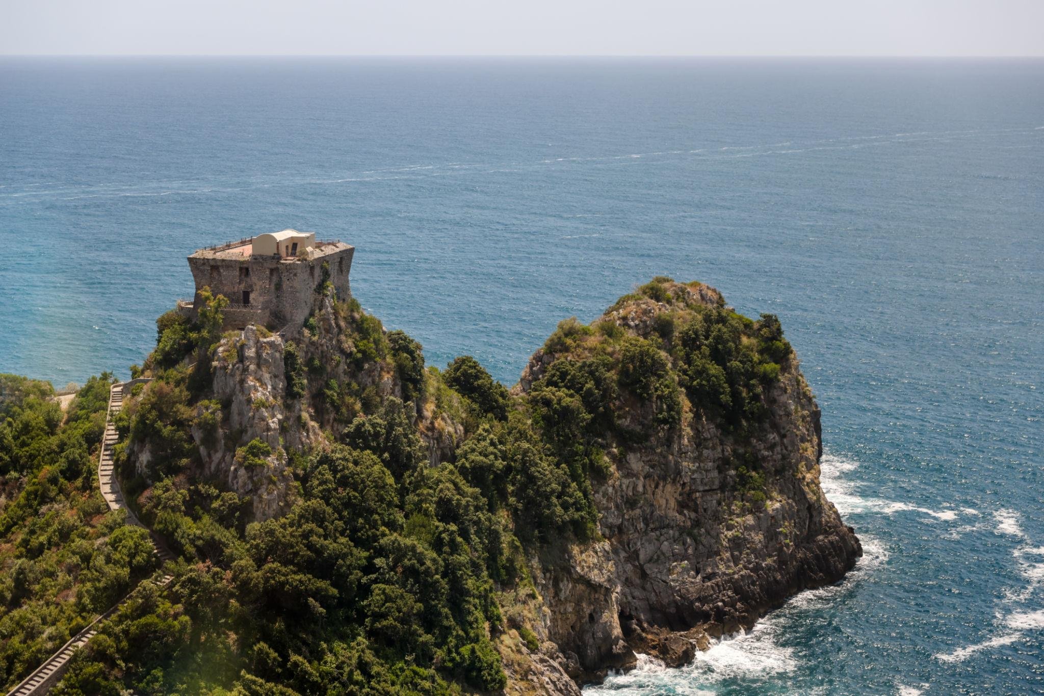 A historic stone watchtower on a rocky cliff overlooking the ocean, with a staircase winding through lush greenery at the bottom.