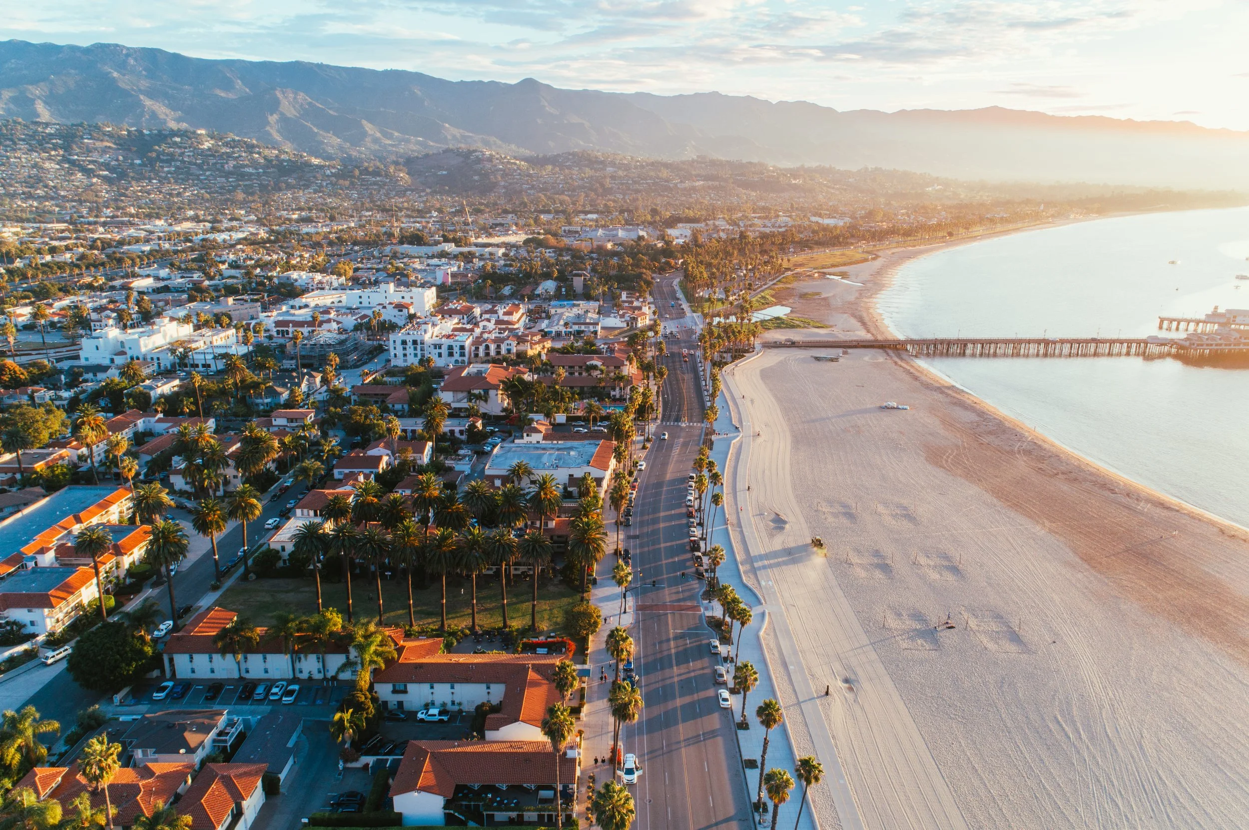 An aerial view of a coastal city with houses and tall palm trees along the streets, a sandy beach, and a pier extending into the water. Mountains are visible in the background, and the scene is illuminated by sunlight, suggesting early morning or late afternoon.