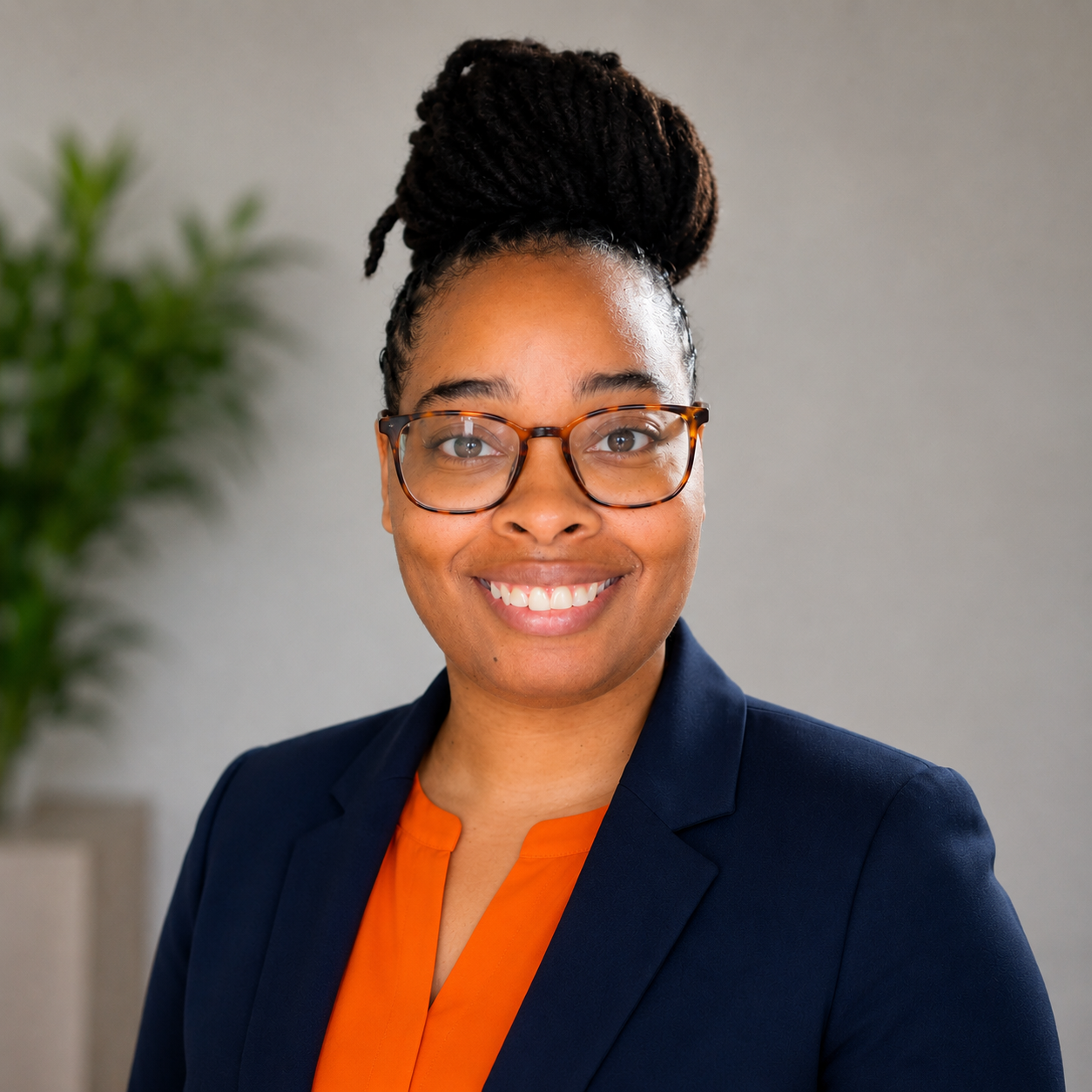 Professional woman with glasses smiling, wearing a navy blazer and orange blouse, standing indoors with a potted plant in the background.