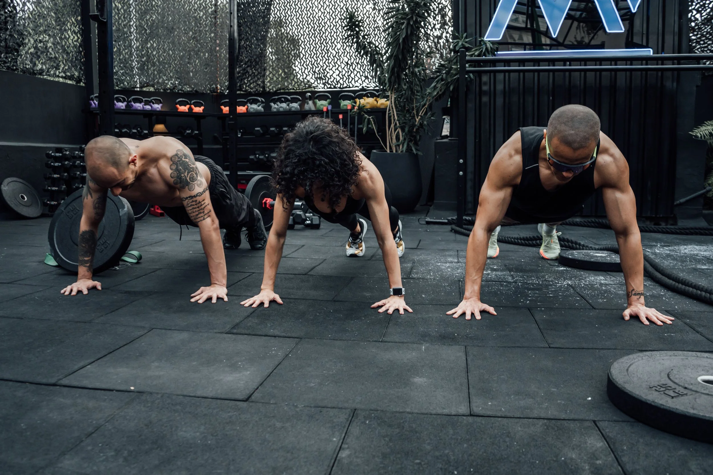 Three people with athletic build and tattoos on their arms doing push-ups in a gym, with weights and kettlebells in the background.