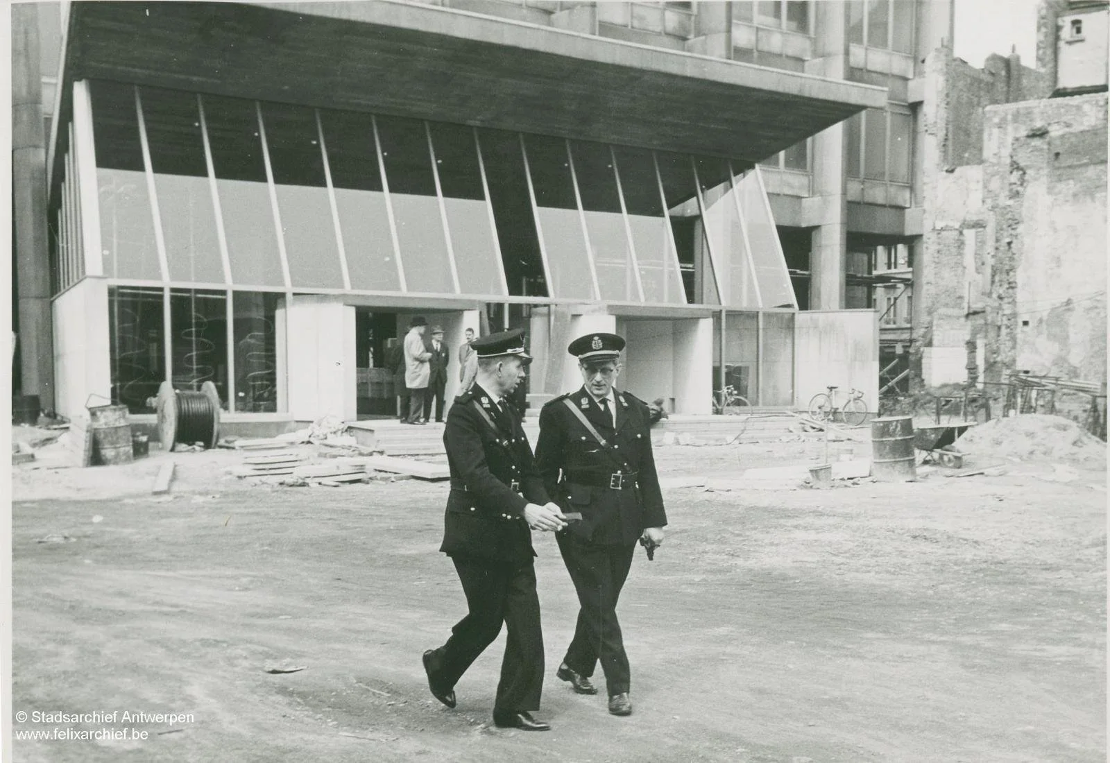 Black and white photo of two police officers walking in front of a modern building under construction, with construction materials and scaffolding visible. In the background, three men are standing near the building.