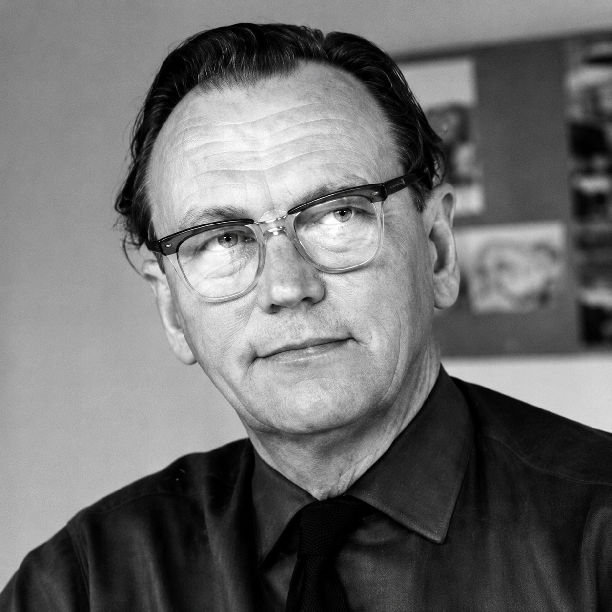 Black and white photo of a man with glasses, wearing a black shirt and tie, looking to the side with a serious expression.