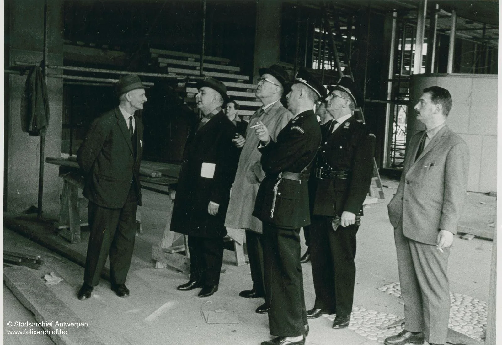 A group of men, including police officers and civilians, standing in a line inside a building under construction, with scaffolding and construction materials visible in the background.