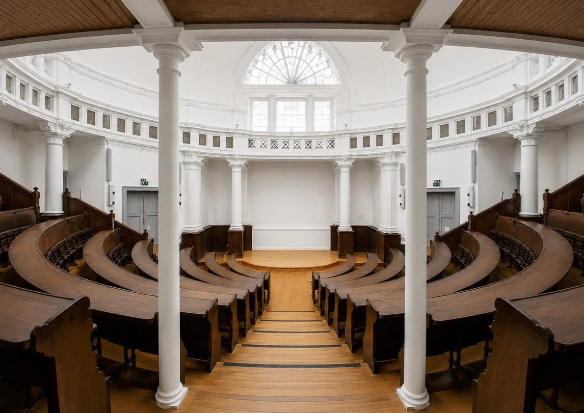 Empty lecture hall with wooden desks, white columns, and large circular window