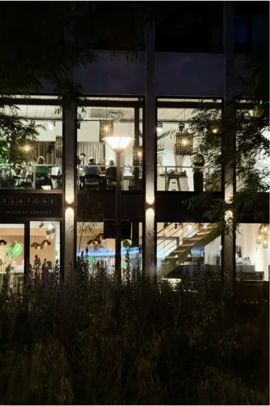 Night view of a two-story restaurant with large windows, interior lighting, and visible plant decor, viewed from outside through trees and bushes.