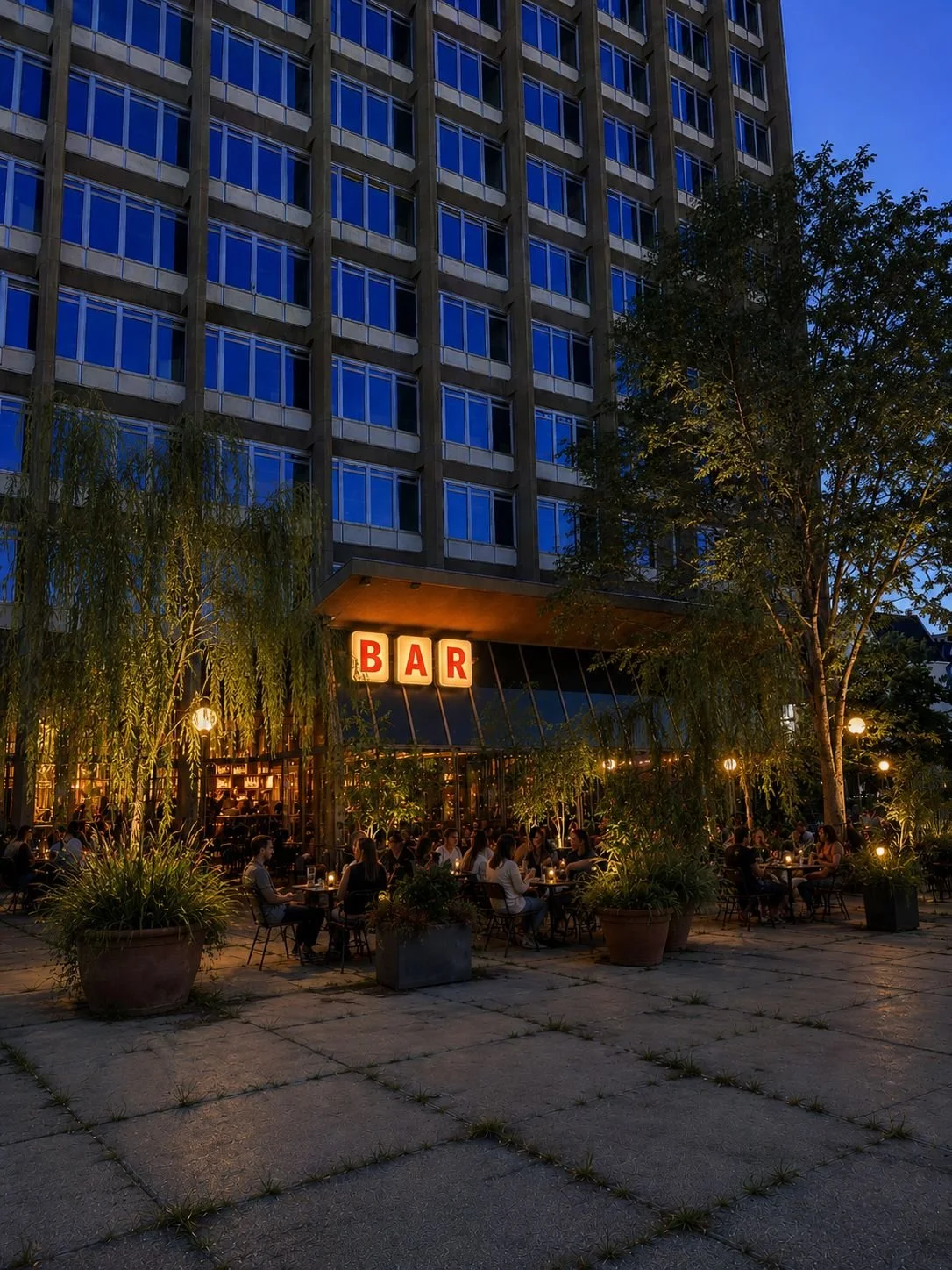 A lively outdoor bar and restaurant area at dusk with patrons seated at tables, potted plants, trees, and warm string lights, located beneath a multi-story building with blue-tinted windows.