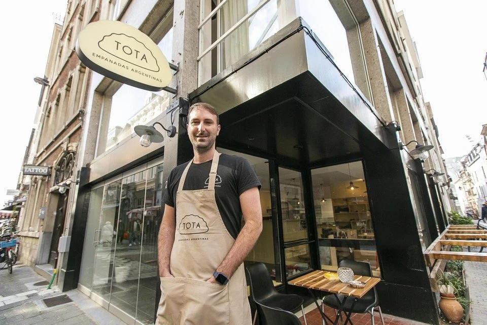 A man standing outside a restaurant called TOTA Empanadas Argentinas, wearing a black t-shirt and beige apron, smiling at the camera.