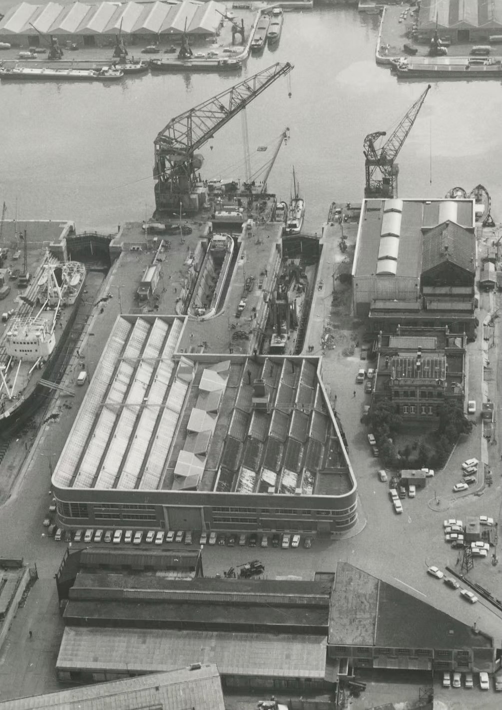 Aerial view of a harbor with boats, cranes, and buildings, including a large modern building with a curved roof and parked cars.