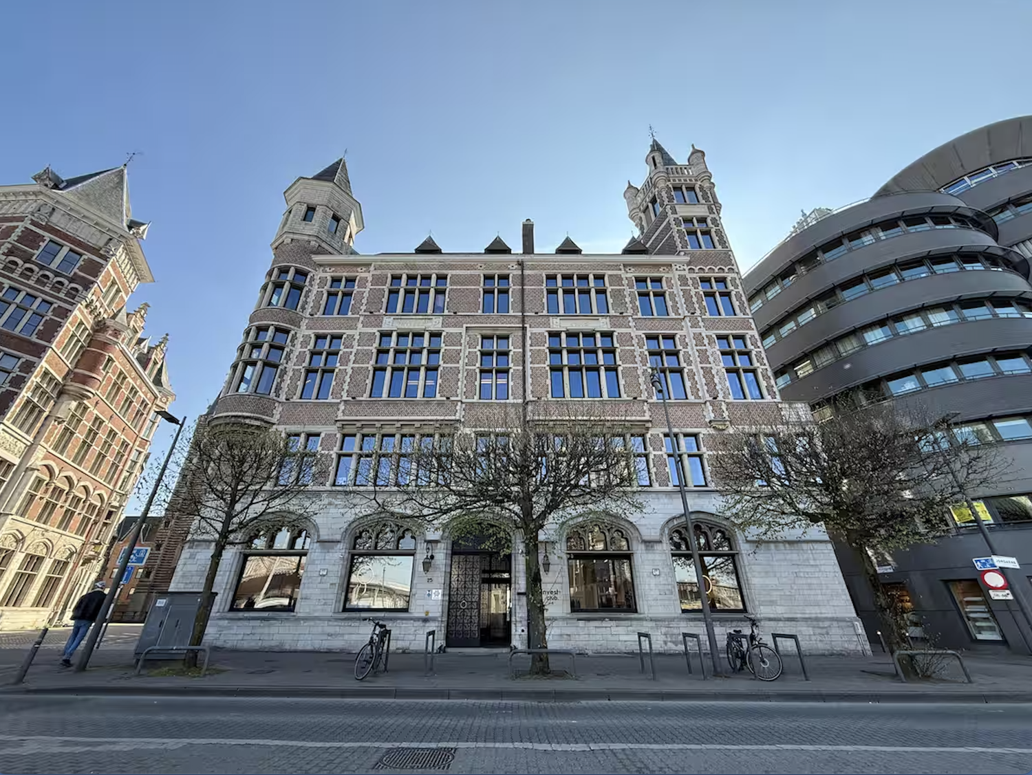 A historic multi-story brick and stone building with ornate architecture, surrounded by leafless trees, bicycles, and modern buildings, under a clear blue sky.