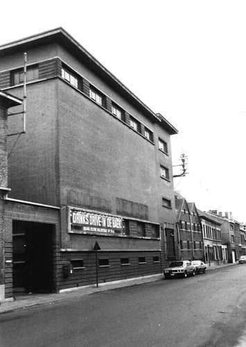 Black and white photo of a multi-story building on a city street with parked cars, and a sign in French that says 'Drugs Store de Dube'.
