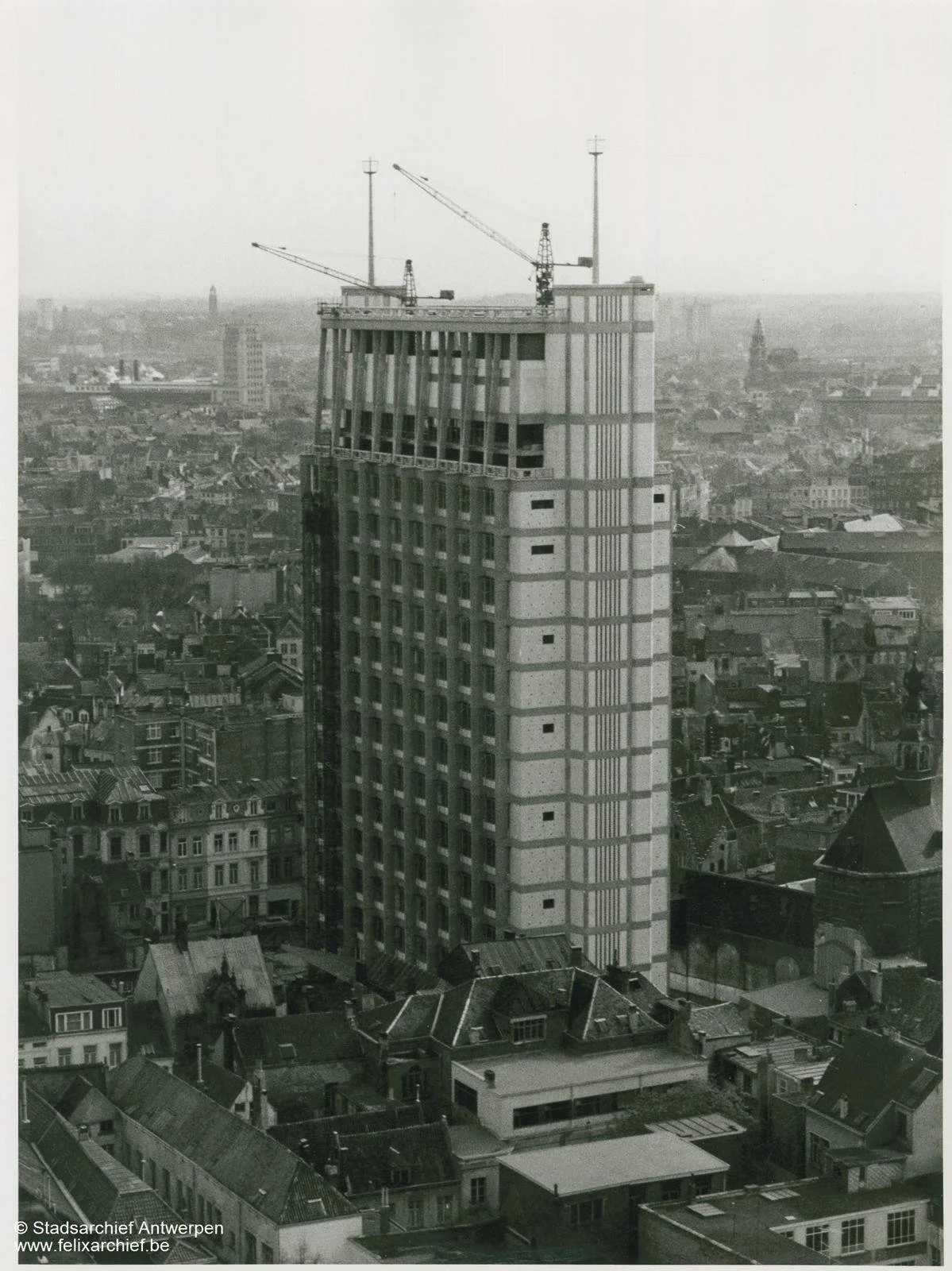 Black and white photo of a tall building under construction, with cranes on top, overlooking a dense cityscape with various rooftops.