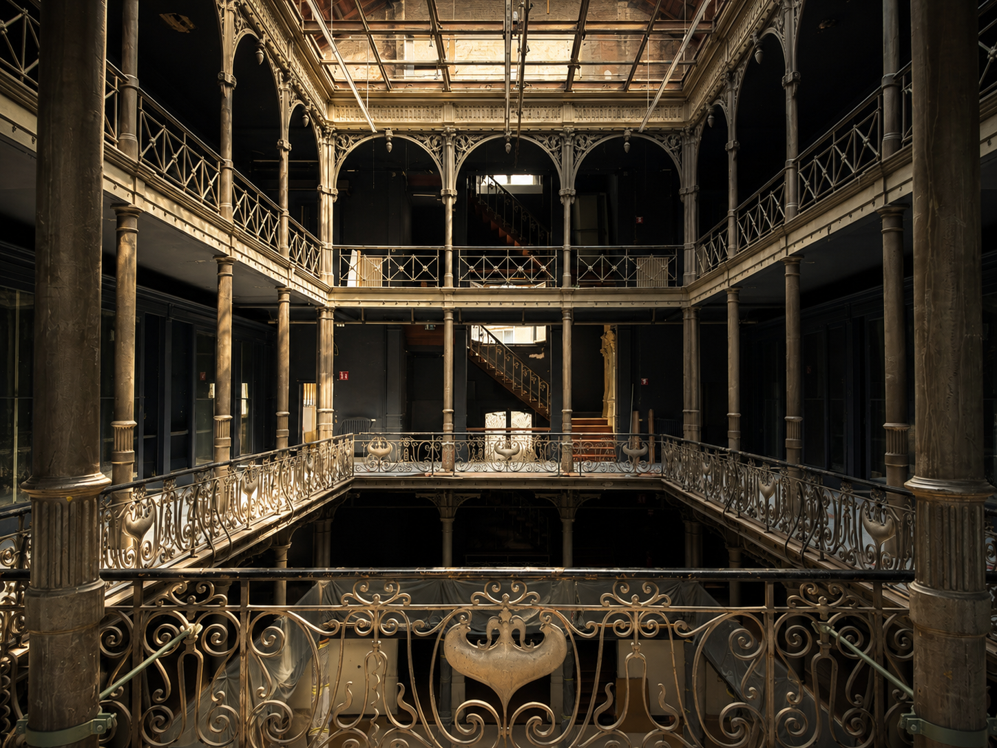 Interior view of an ornate, multi-level building with decorative iron railings, columns, and stairs, lit by natural light from a skylight.