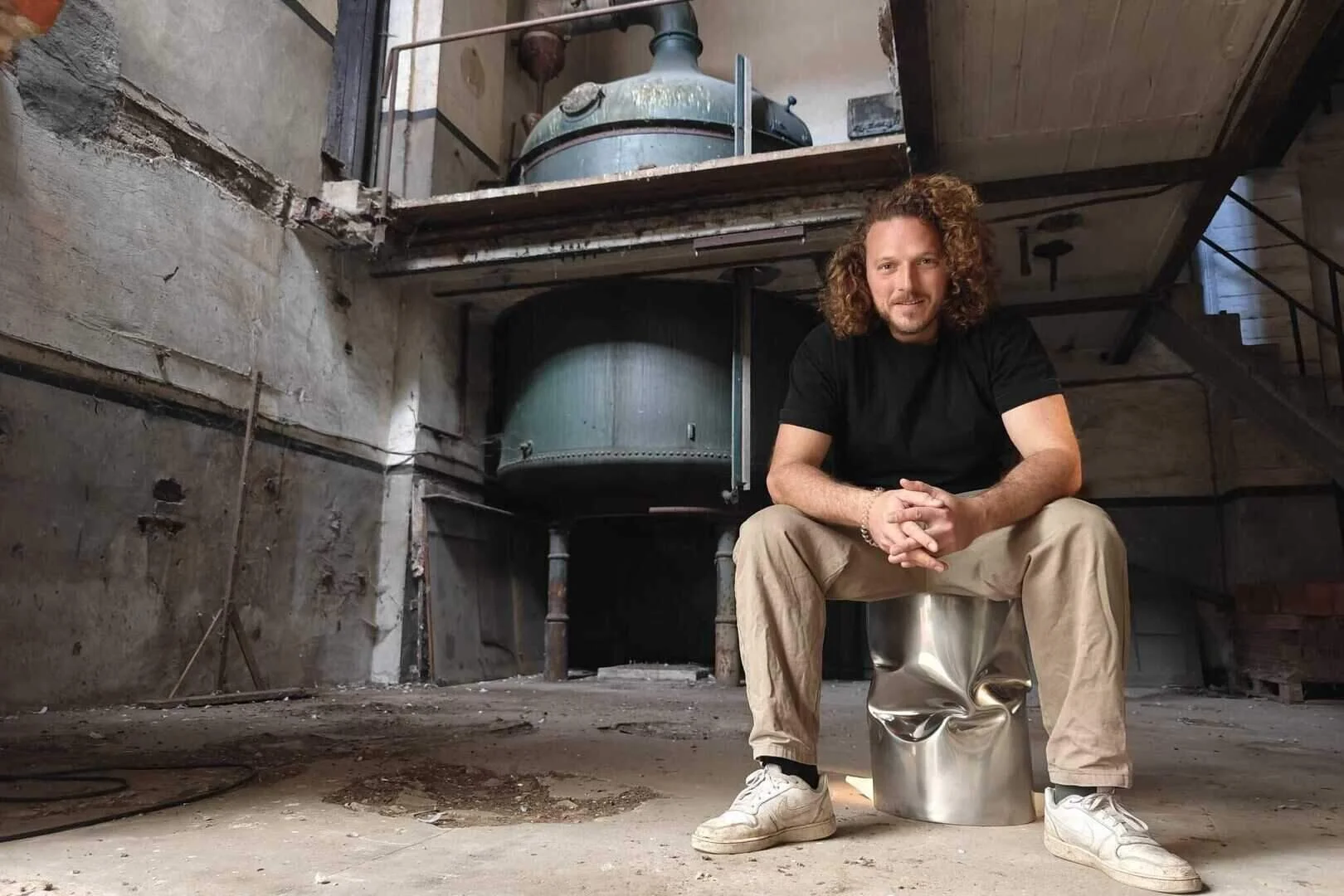 A man with curly hair and a beard, wearing a black T-shirt and beige pants, sitting on a metallic stool in an industrial, unfinished space with concrete walls, a large industrial kettle, and a staircase.