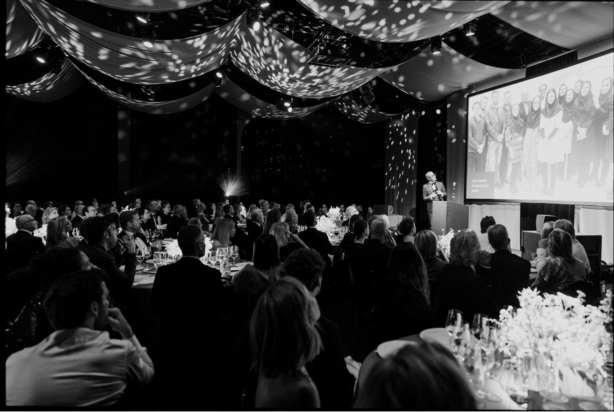 A large formal event with many attendees sitting at round tables, watching a speaker on stage. The stage has a large screen displaying a group photo of people. Decorated ceiling with draped fabric and constellation-like lighting patterns.