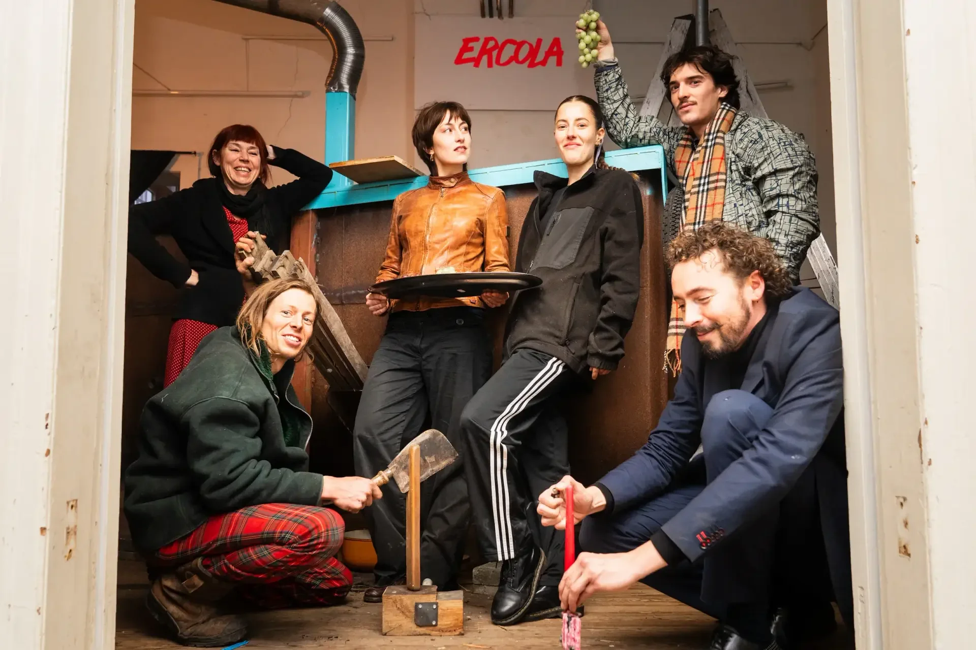 A group of seven people posing in a small, cluttered room with a white wall and a sign that says 'ERCOLA' with a bunch of grapes hanging from their hand. Four people are standing, two are crouching, and one is sitting. Some are holding tools like a hammer and an anvil, and they are all dressed casually.