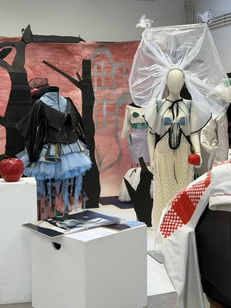 Display of avant-garde fashion outfits featuring a mannequin with large, transparent headpiece, colorful and layered clothing, and artistic backdrop with painted tree and house, in a styled boutique or art installation.