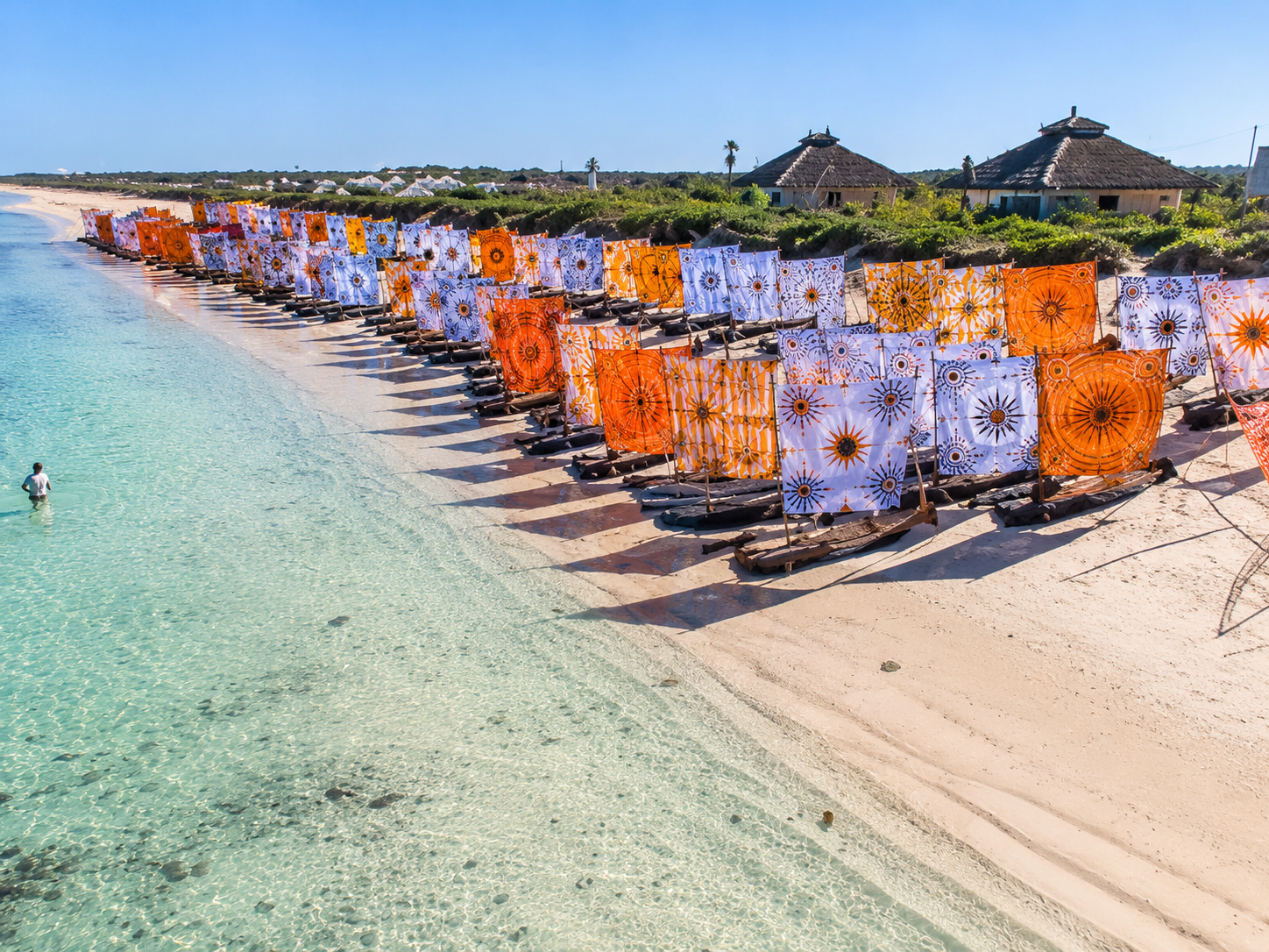 Colorful patterned beach umbrellas on a sandy beach with calm, clear water and a person swimming near the shore, with thatched roof huts and greenery in the background.