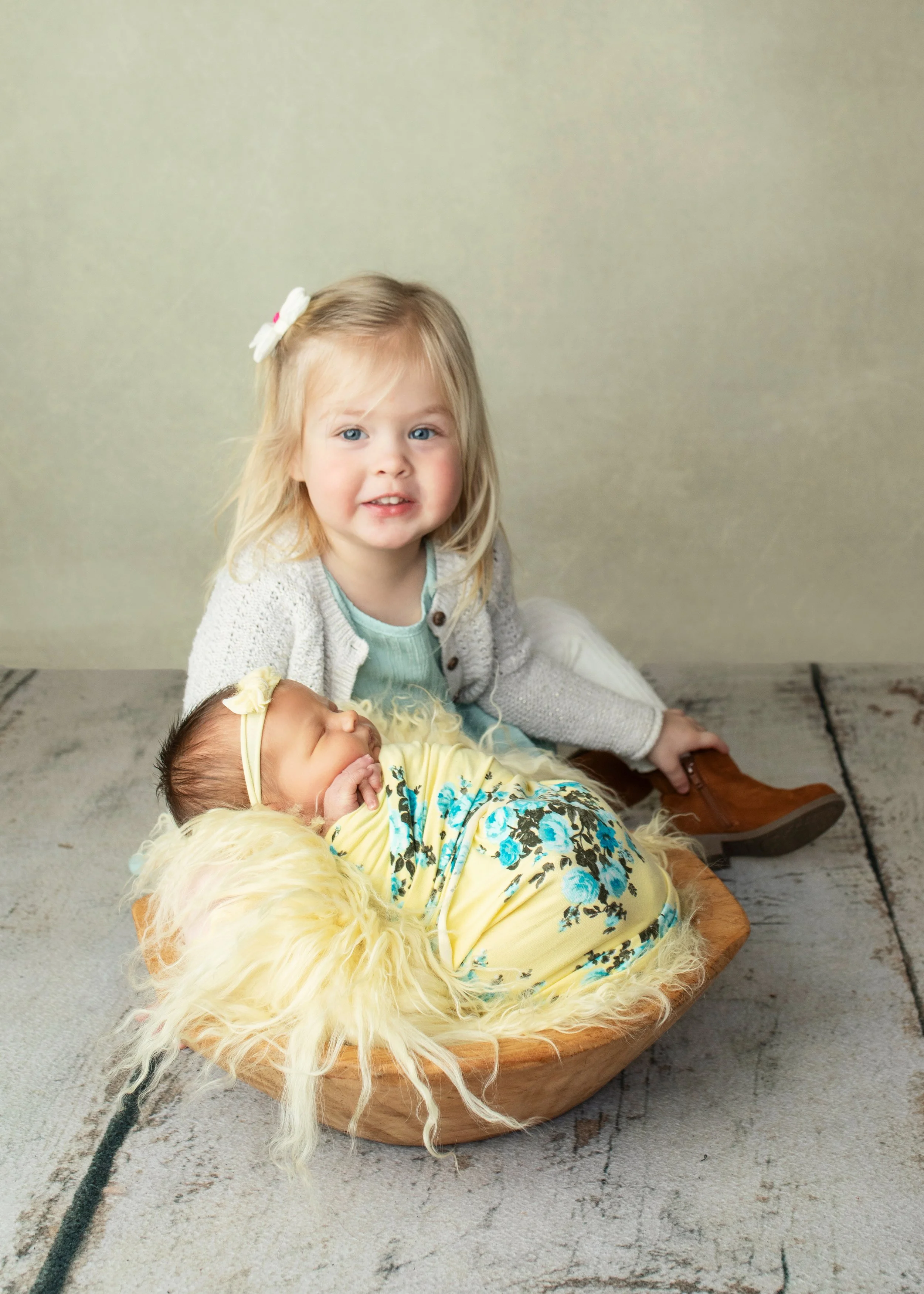 A young girl with blonde hair, blue eyes, and a white bow in her hair, sits on the floor next to a newborn baby in a wooden bowl. The baby is wearing a yellow floral dress, a cream-colored jacket, and a yellow headband. The girl is dressed in a light blue shirt and a gray cardigan, and is smiling at the camera against a plain beige background.