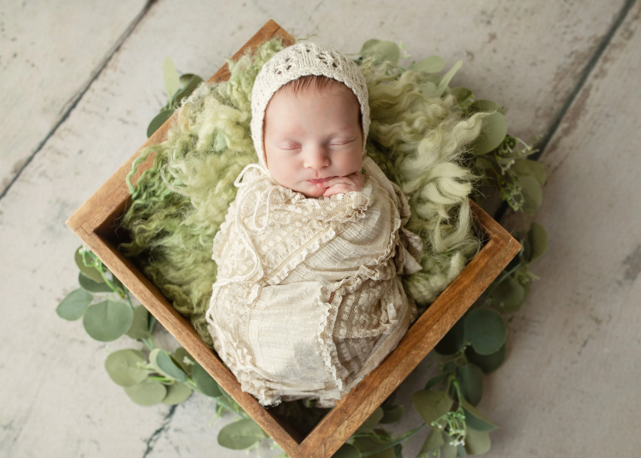 A sleeping baby lying in a wooden box with green fluffy blanket, wearing a beige lace dress and knitted cream hat, surrounded by green eucalyptus leaves.