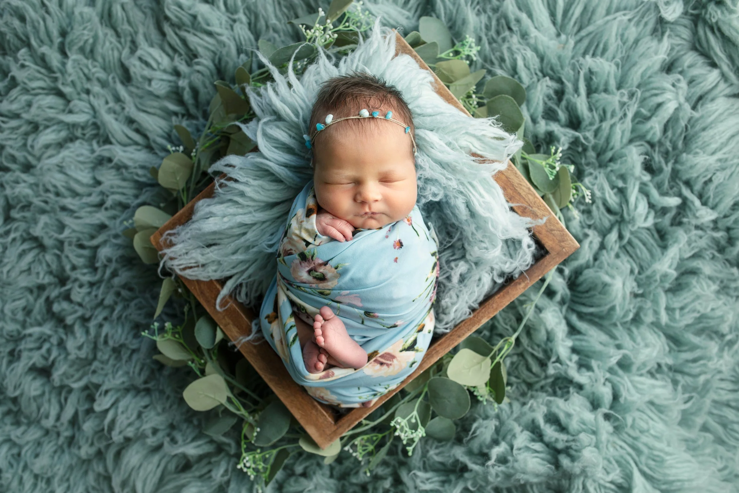 A sleeping baby with a floral headband, wrapped in a floral blanket, lying on a fluffy green rug, in a wooden basket with eucalyptus leaves and white flowers around.