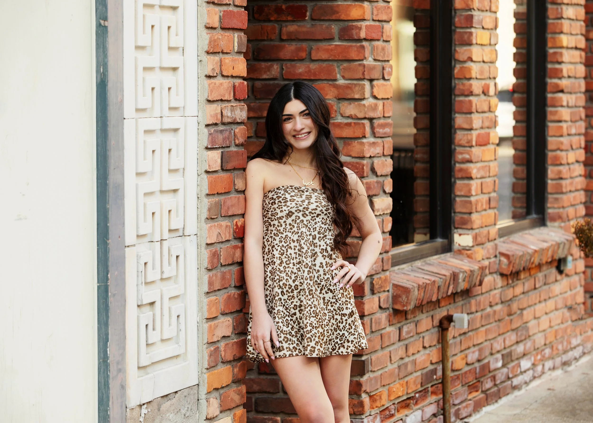 A woman with dark hair smiling and leaning against a red brick wall, wearing a leopard print dress.