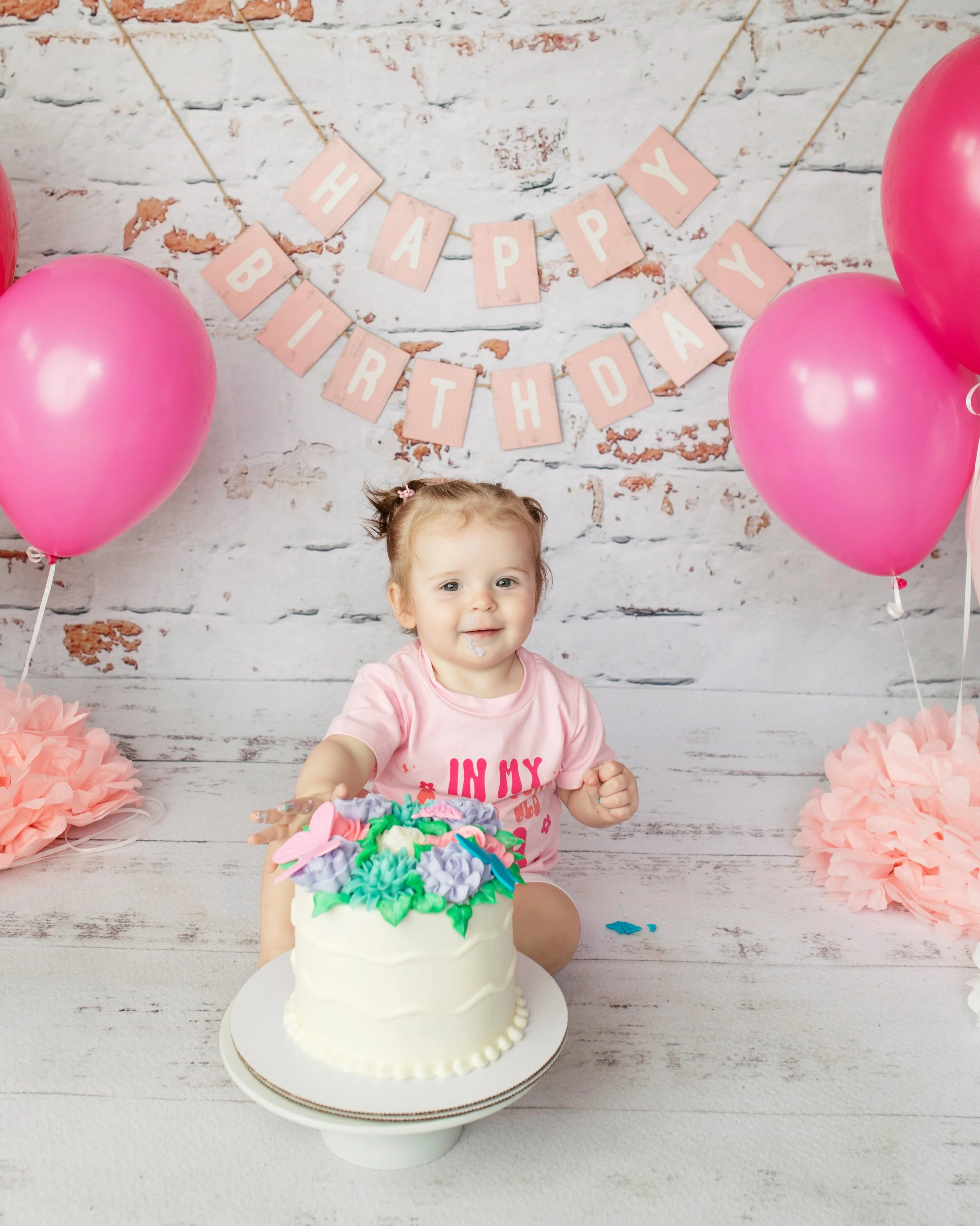 A young girl with a pink shirt celebrating her birthday at a party with pink balloons, a decorative banner that says 'Happy Birthday', and a white cake with colorful frosting and floral decorations.