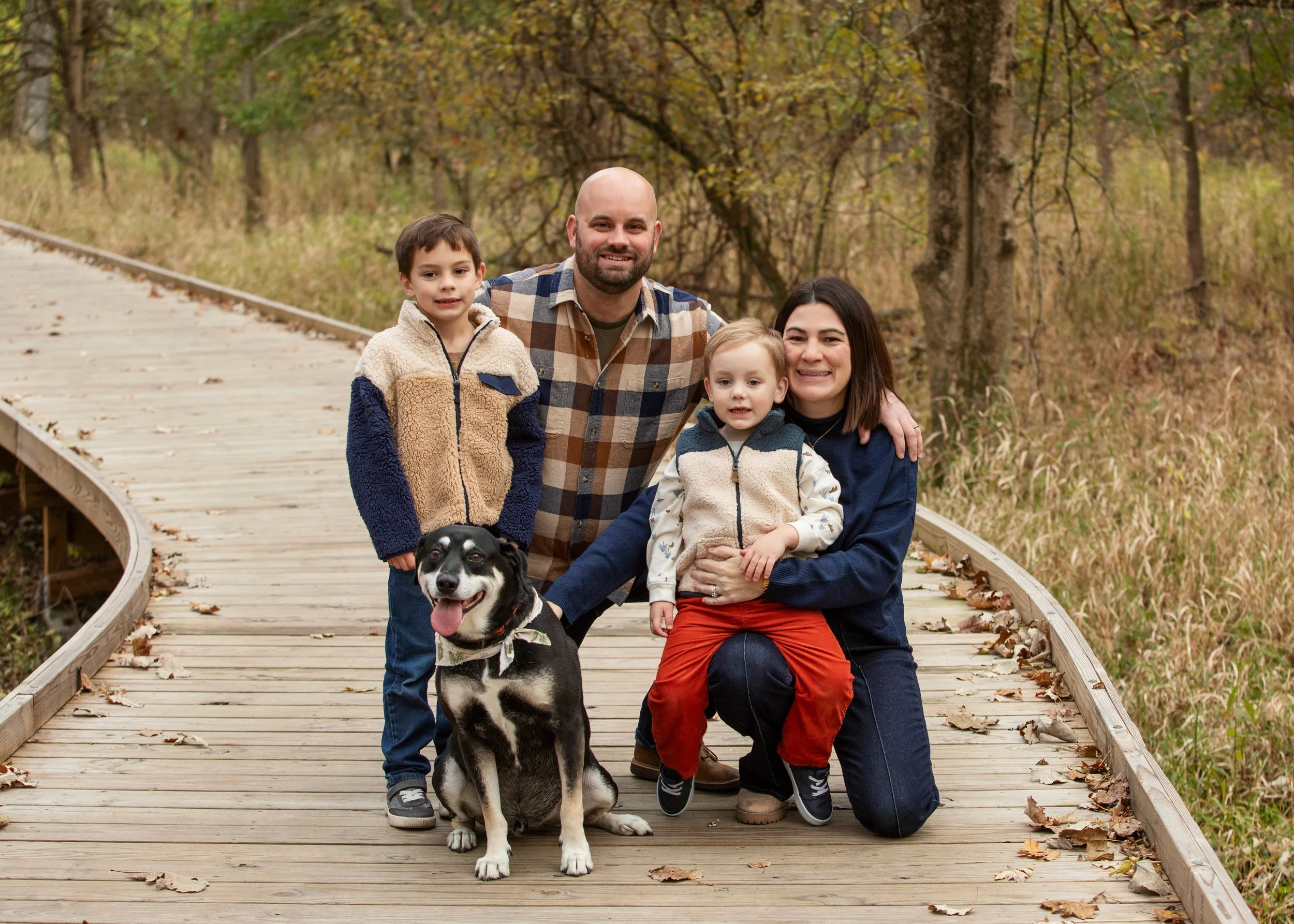 A family of four with two children, an adult man and an adult woman, along with a black and white dog, standing on a wooden bridge in a wooded area during fall. The family is smiling at the camera.