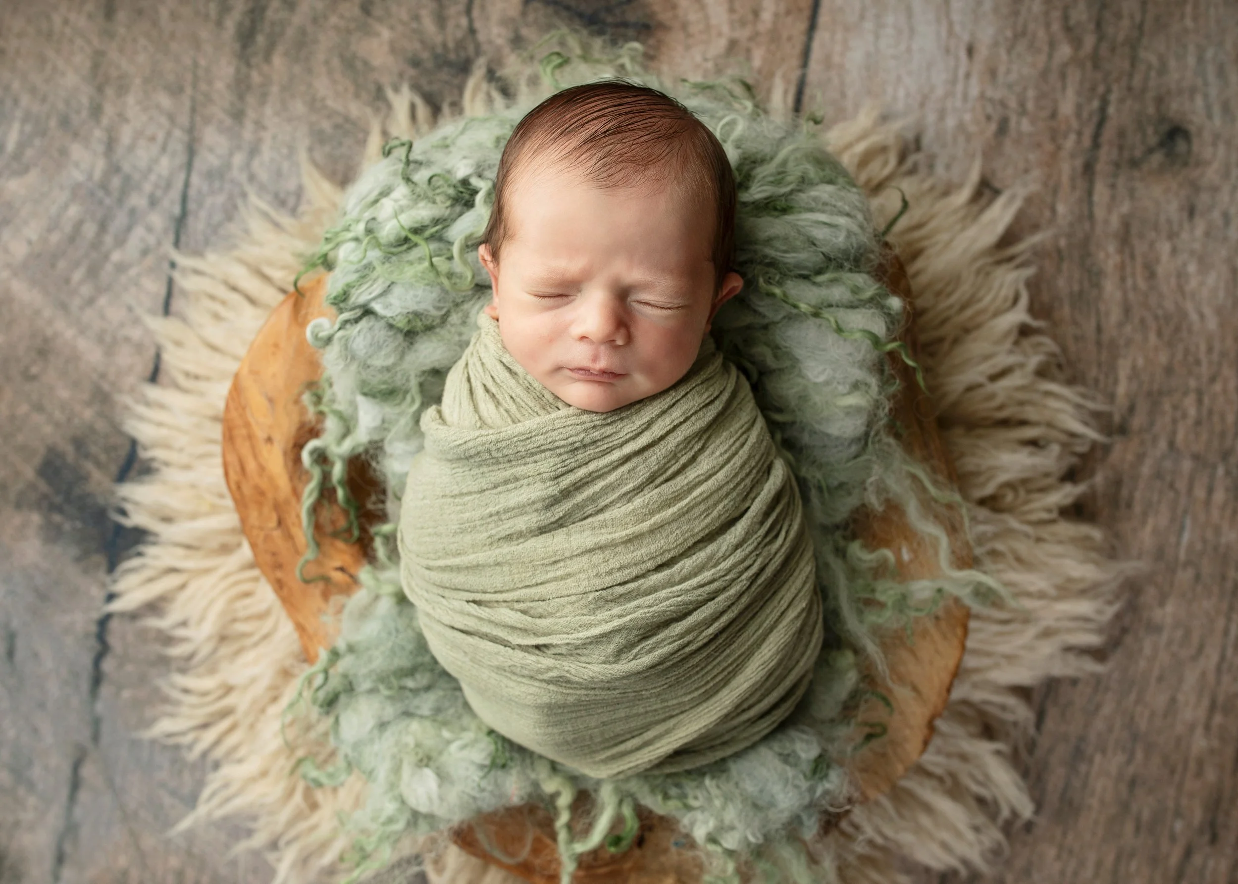 A newborn baby with closed eyes wrapped in a green cloth, lying on a fluffy cream-colored rug with a textured background.
