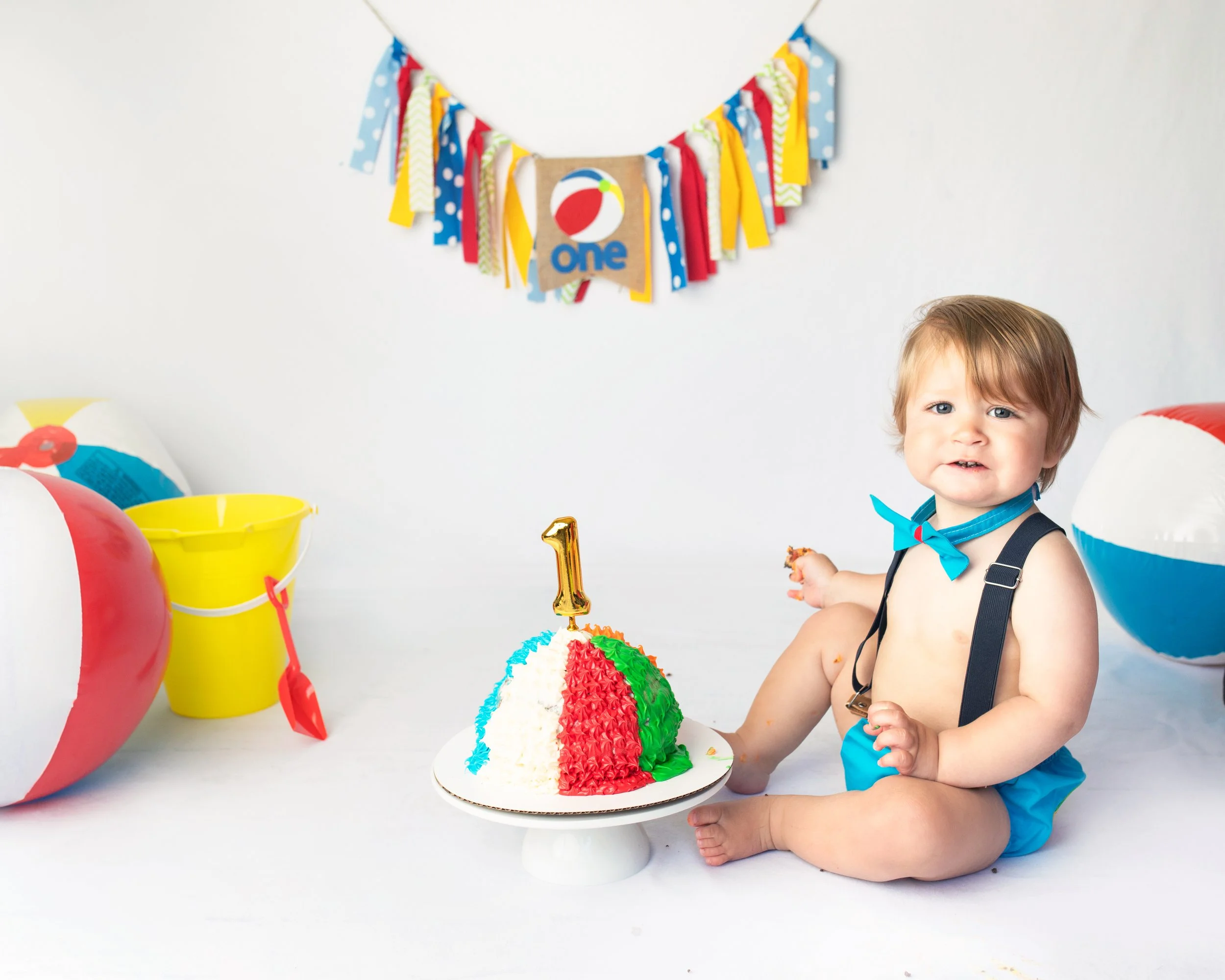 Young child with light brown hair and blue eyes, wearing a blue outfit with suspenders and a blue bow tie, celebrating a birthday with a colorful cake, balloons, a birthday banner, and a gold number one candle on the cake.