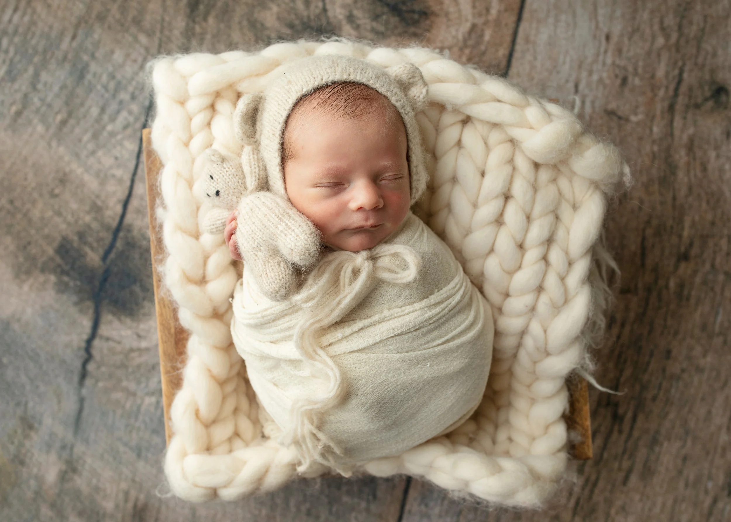 A sleeping baby wearing a knitted hat with bear ears, swaddled in a cream-colored blanket, and lying on a chunky knit blanket. The baby is on a wooden surface with a rustic, textured background.