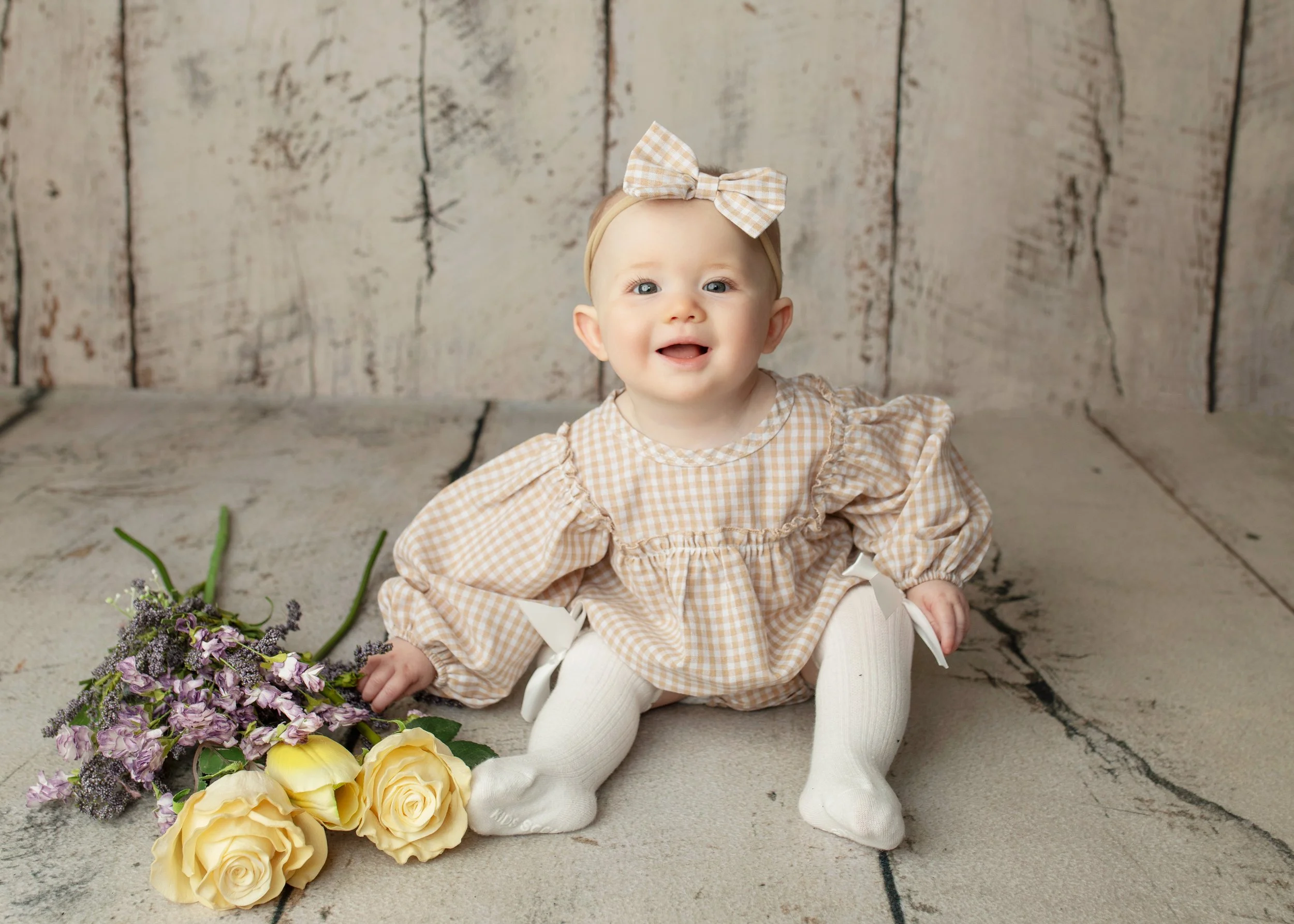 A baby girl with blue eyes, wearing a beige headband with a checkered bow and a matching dress, sitting on the floor near a bouquet of purple and yellow flowers against a rustic wooden background.