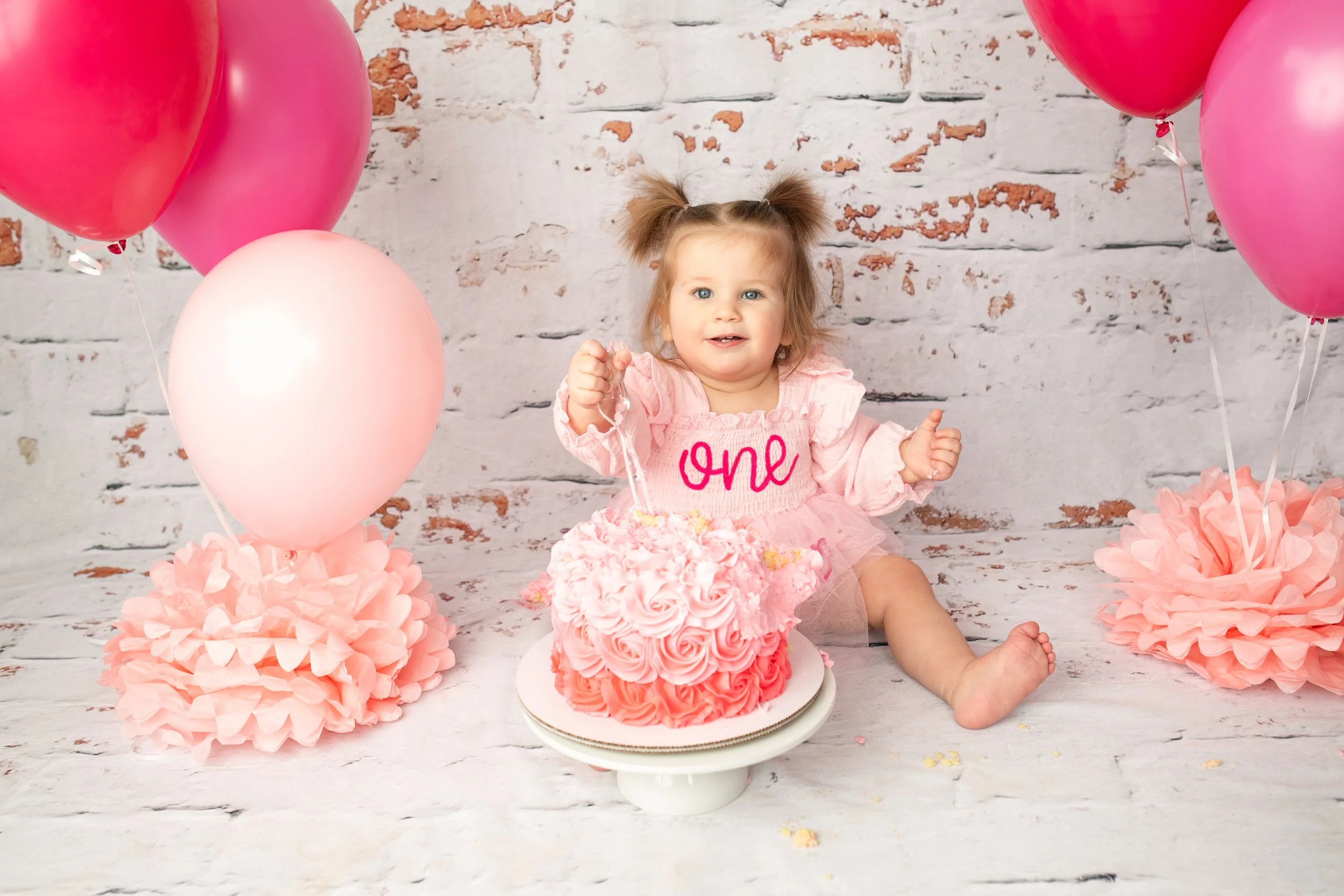 A young girl celebrating her first birthday with pink balloons, a pink cake with floral decoration, and a "one" cake topper. She is sitting on the floor in front of a white brick wall, wearing a pink outfit.