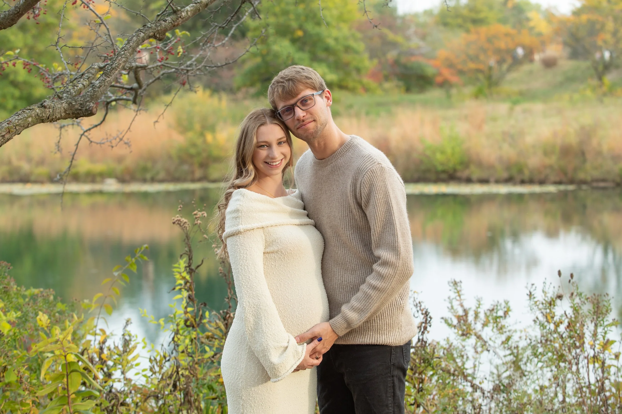A young woman and a young man standing close together outdoors by a lake with autumn foliage in the background. The woman is smiling, wearing a cream dress and wrapped in a blanket, while the man is wearing glasses and a beige sweater.