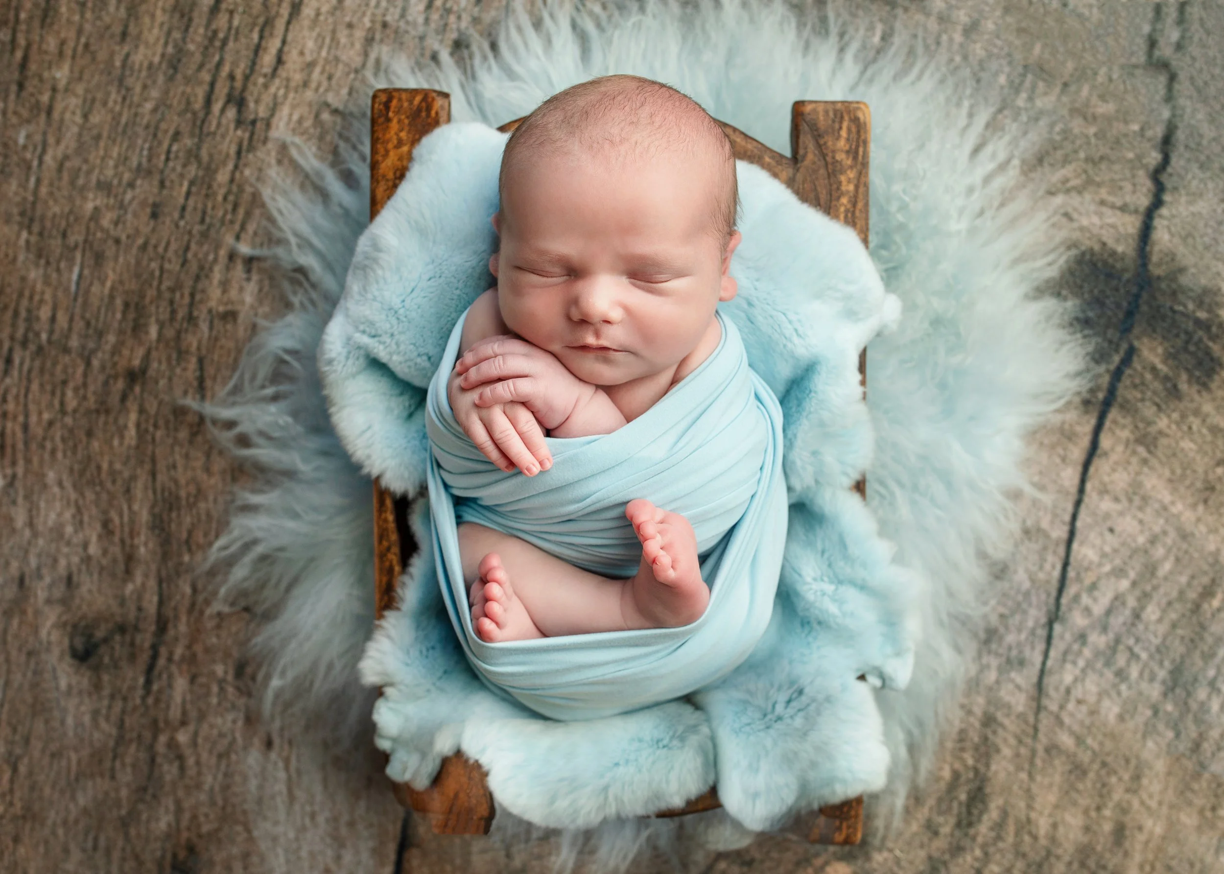 A newborn baby curled up sleeping on a small wooden bed, wrapped in a light blue swaddle, resting on a soft white blanket.