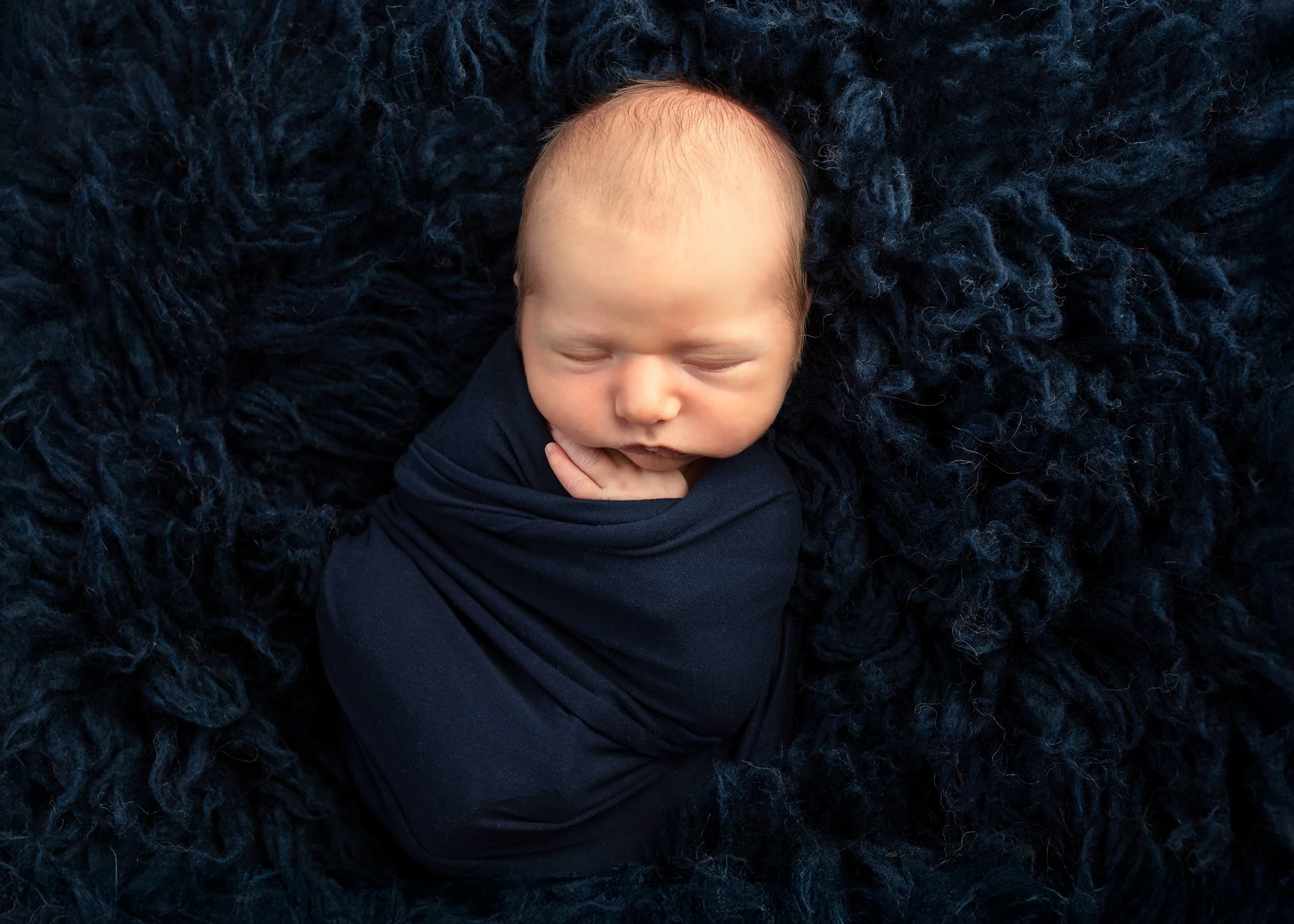Close-up of a sleeping baby lying on a fluffy dark blue blanket, wearing a dark blue outfit.