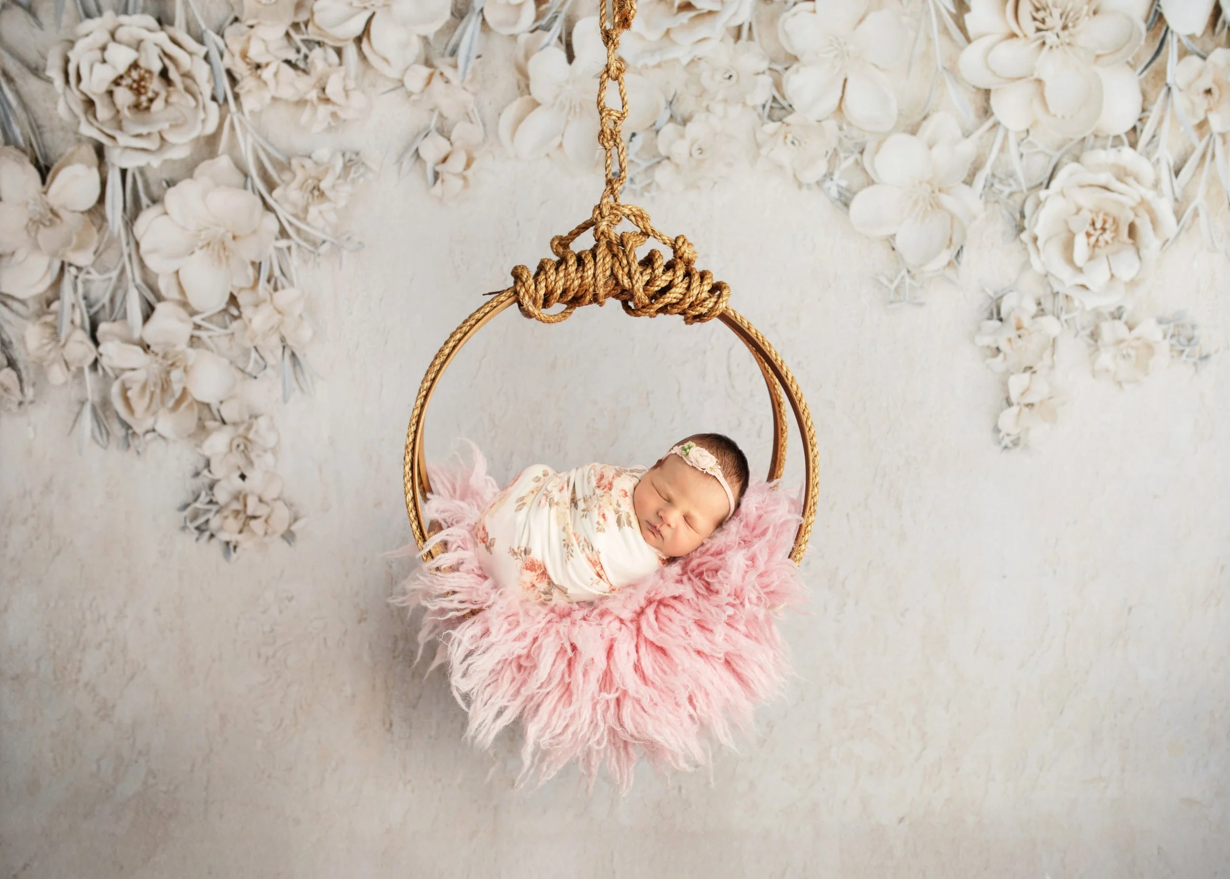 A sleeping baby girl lying on a fluffy pink blanket in a swing, with a floral wallpaper background.