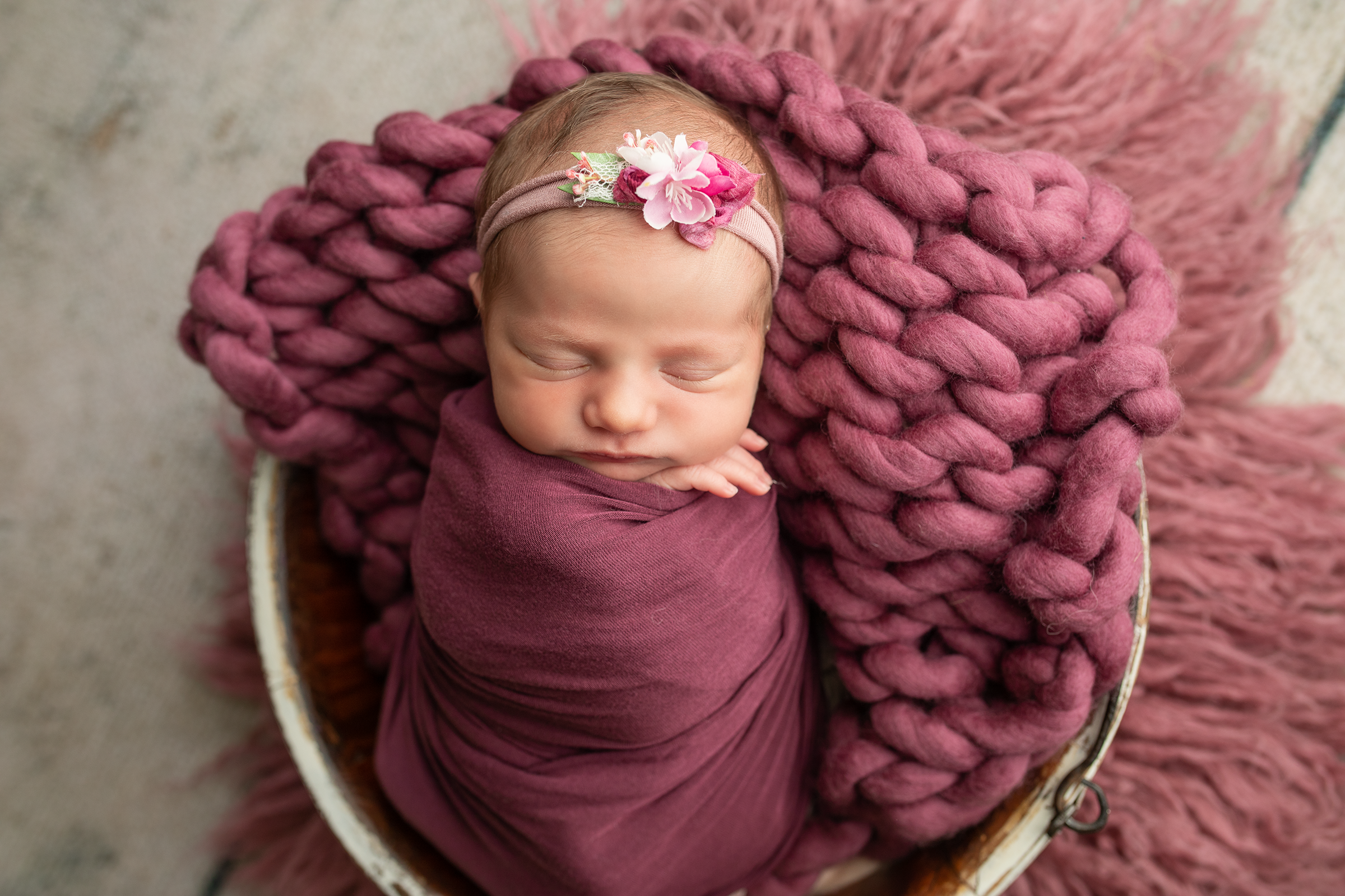 A close-up of a sleeping baby wearing a pink headband with a flower and a pink outfit, lying on a chunky pink knit blanket inside a large bowl or basket.