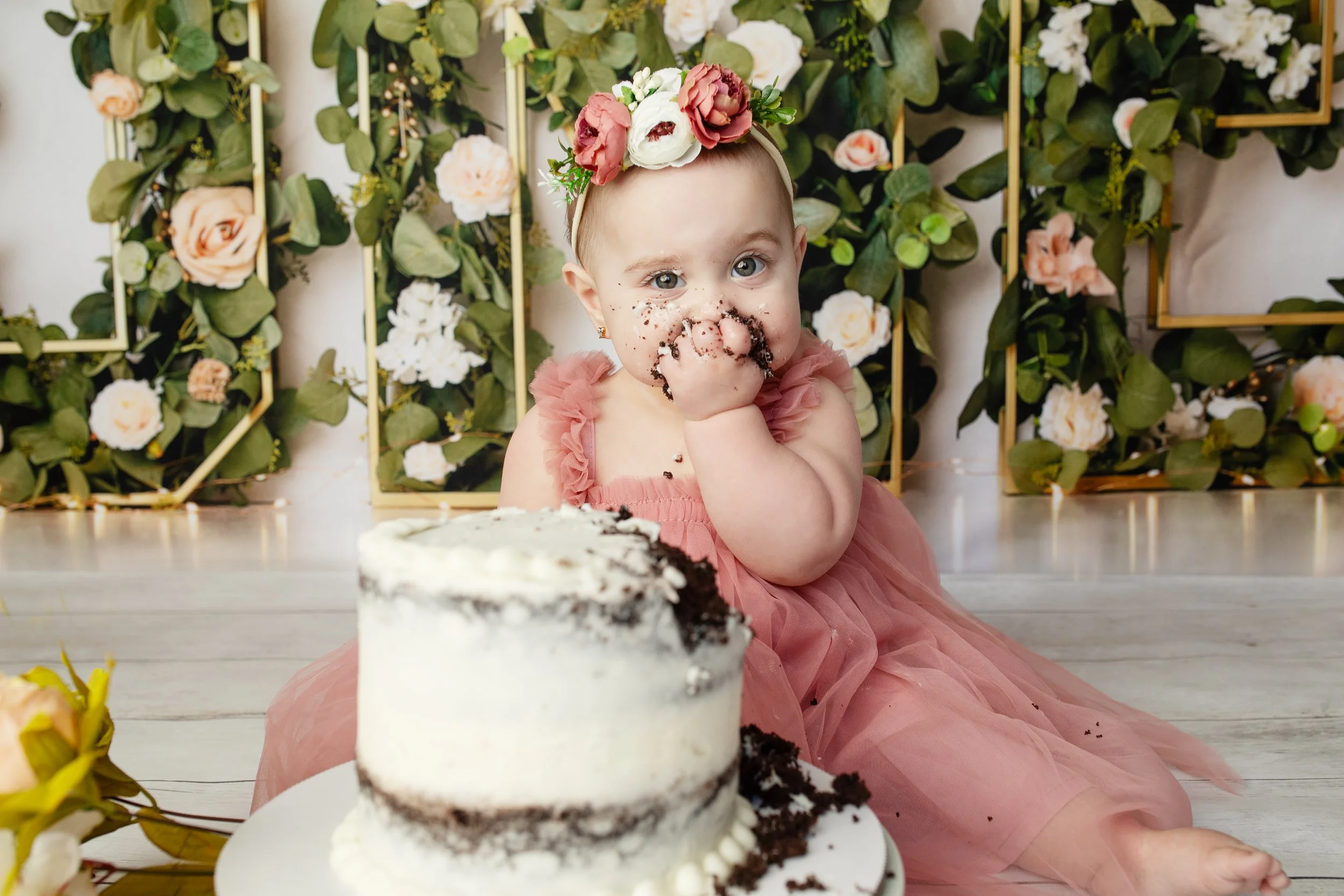 A young girl with big blue eyes and a floral headband, dressed in a pink dress, sitting in front of a decorated backdrop with flowers, enjoying her birthday cake with chocolate crumbs on her face.