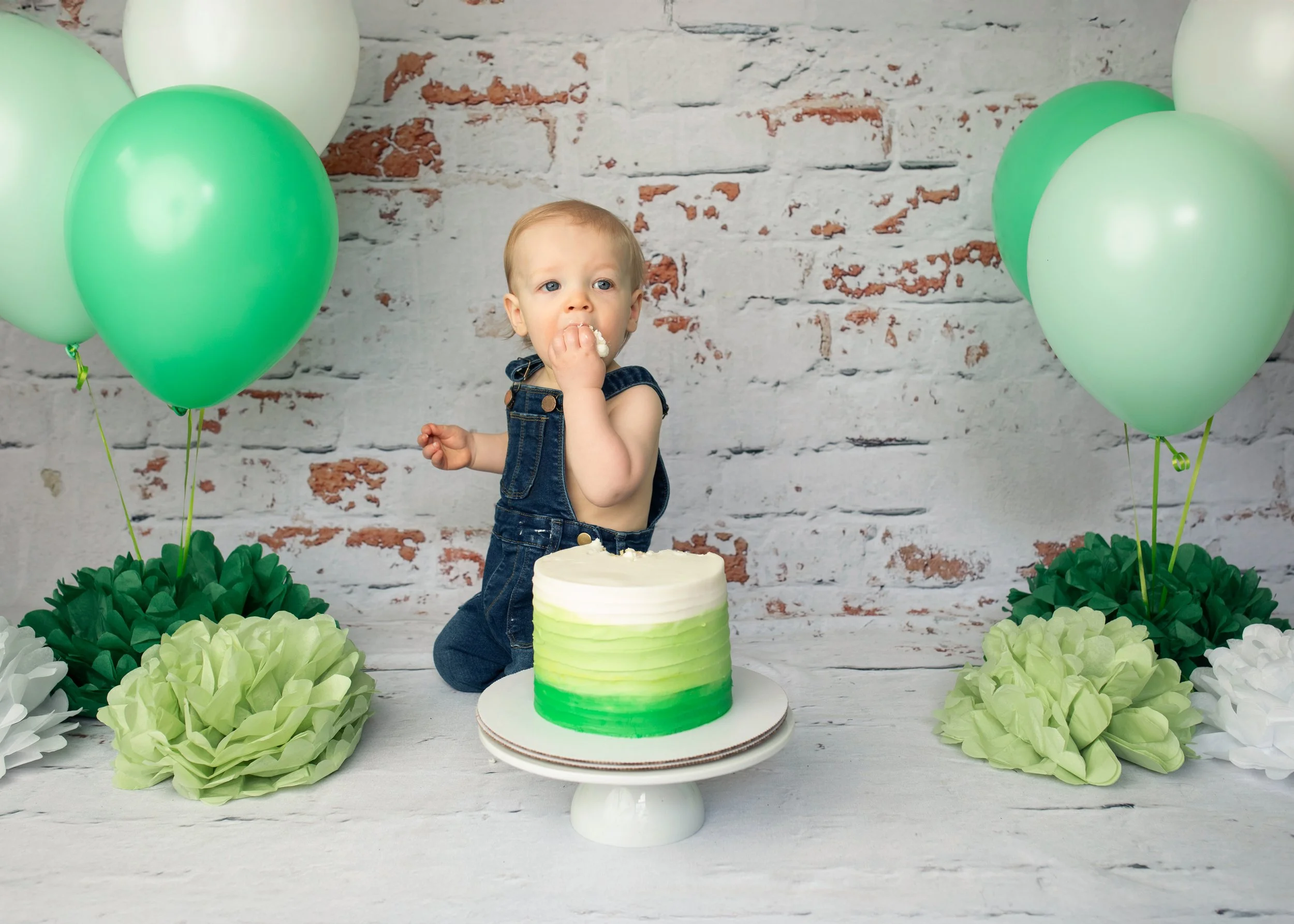 A young child with short blonde hair wearing denim overalls eating cake at a birthday celebration. The cake has a green and white gradient icing. The background features a brick wall with balloons in shades of green and white, and decorative green and white paper flowers surrounding the table.