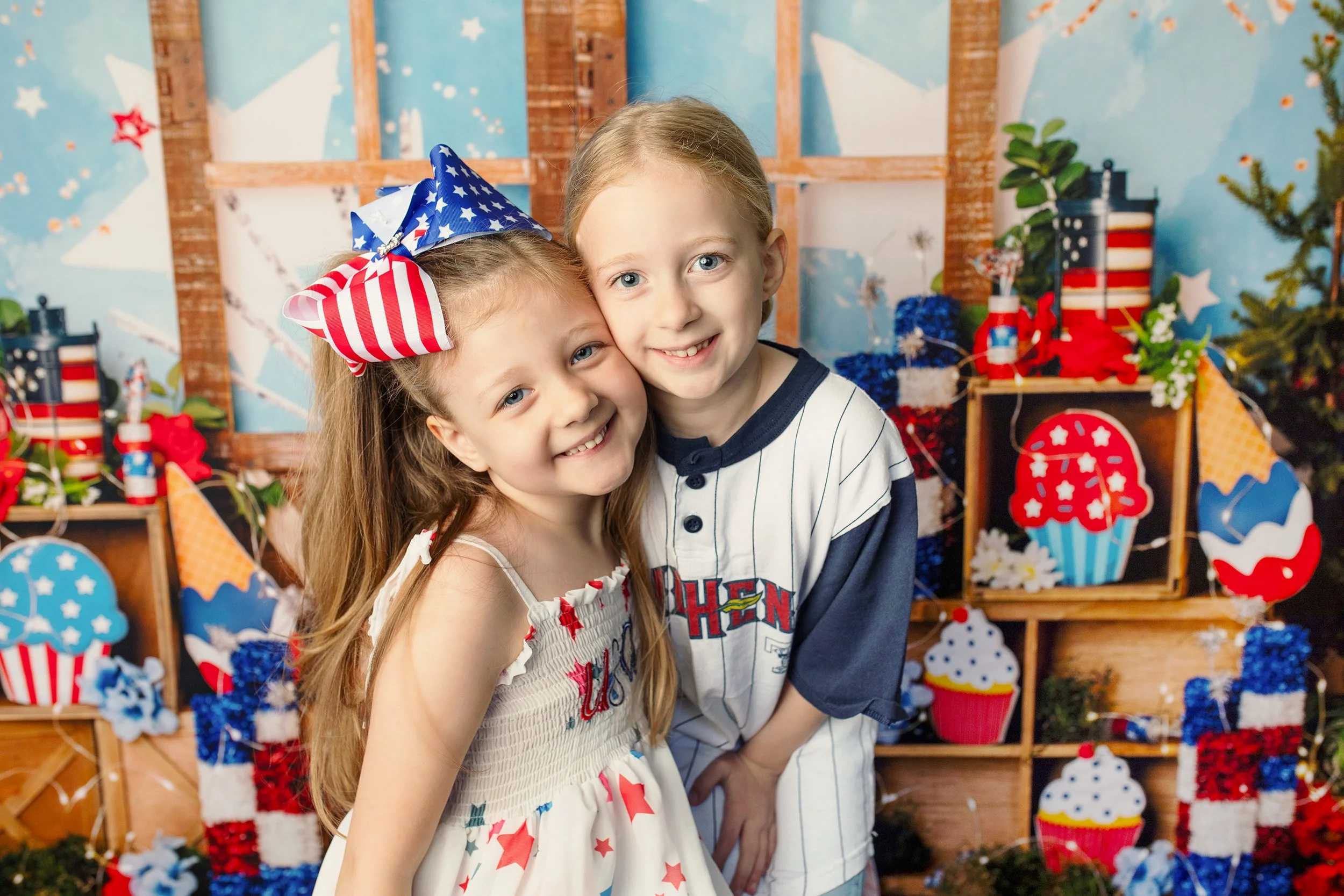 Two young children, a girl and a boy, at a patriotic themed celebration. The girl has long brown hair with a large red, white, and blue bow. The boy has blond hair and blue eyes. They are smiling and hugging each other in front of a background decorated with patriotic items and American flags.