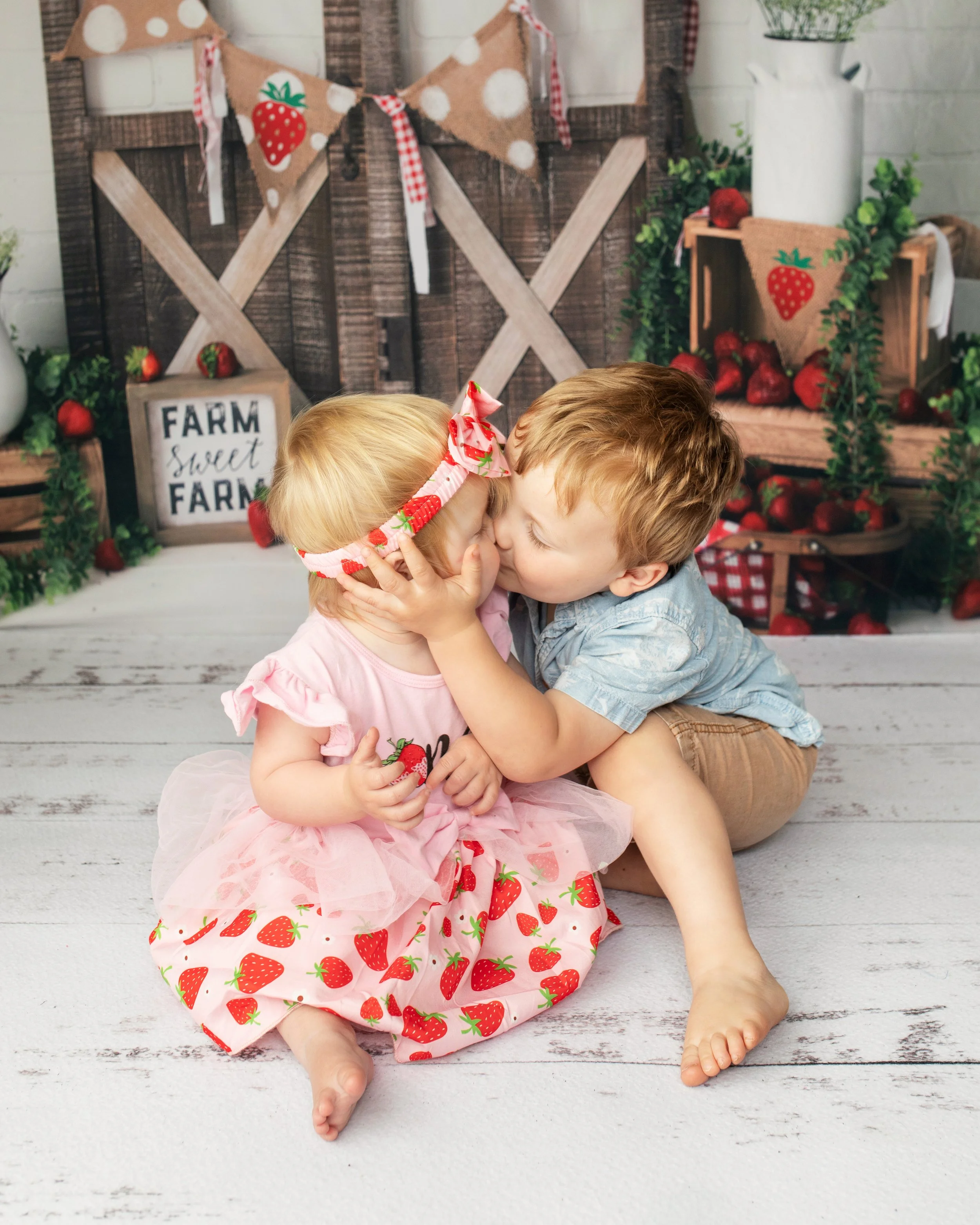 Two young children sharing a kiss or hugging, dressed in strawberry-themed outfits, sitting on a white wooden floor with a farm-themed backdrop decorated with strawberries, greenery, and rustic farm signs.