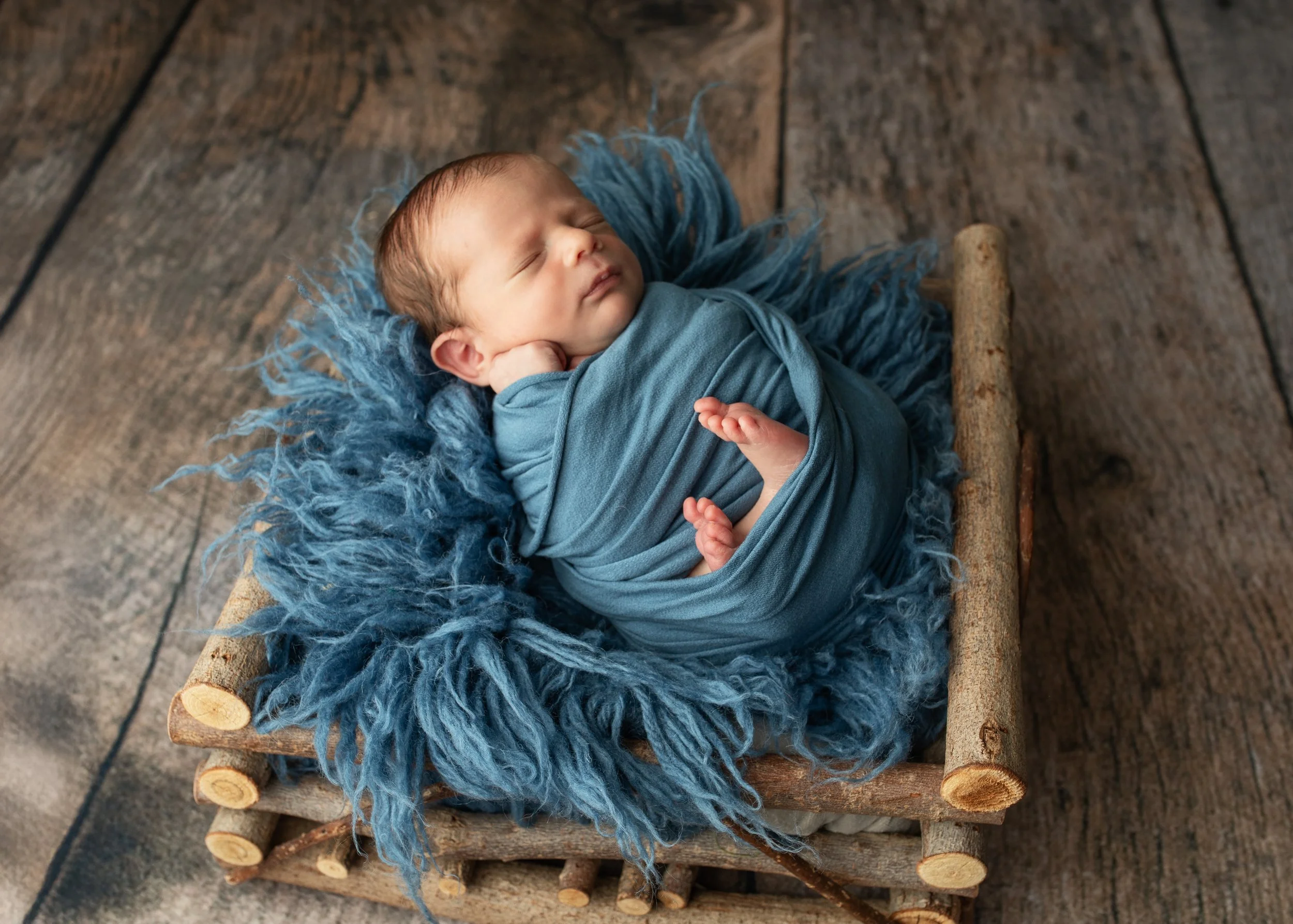 A newborn baby with closed eyes, wrapped in a blue wrap, resting on a furry blue blanket on a small rustic wooden bed, on a wooden floor.