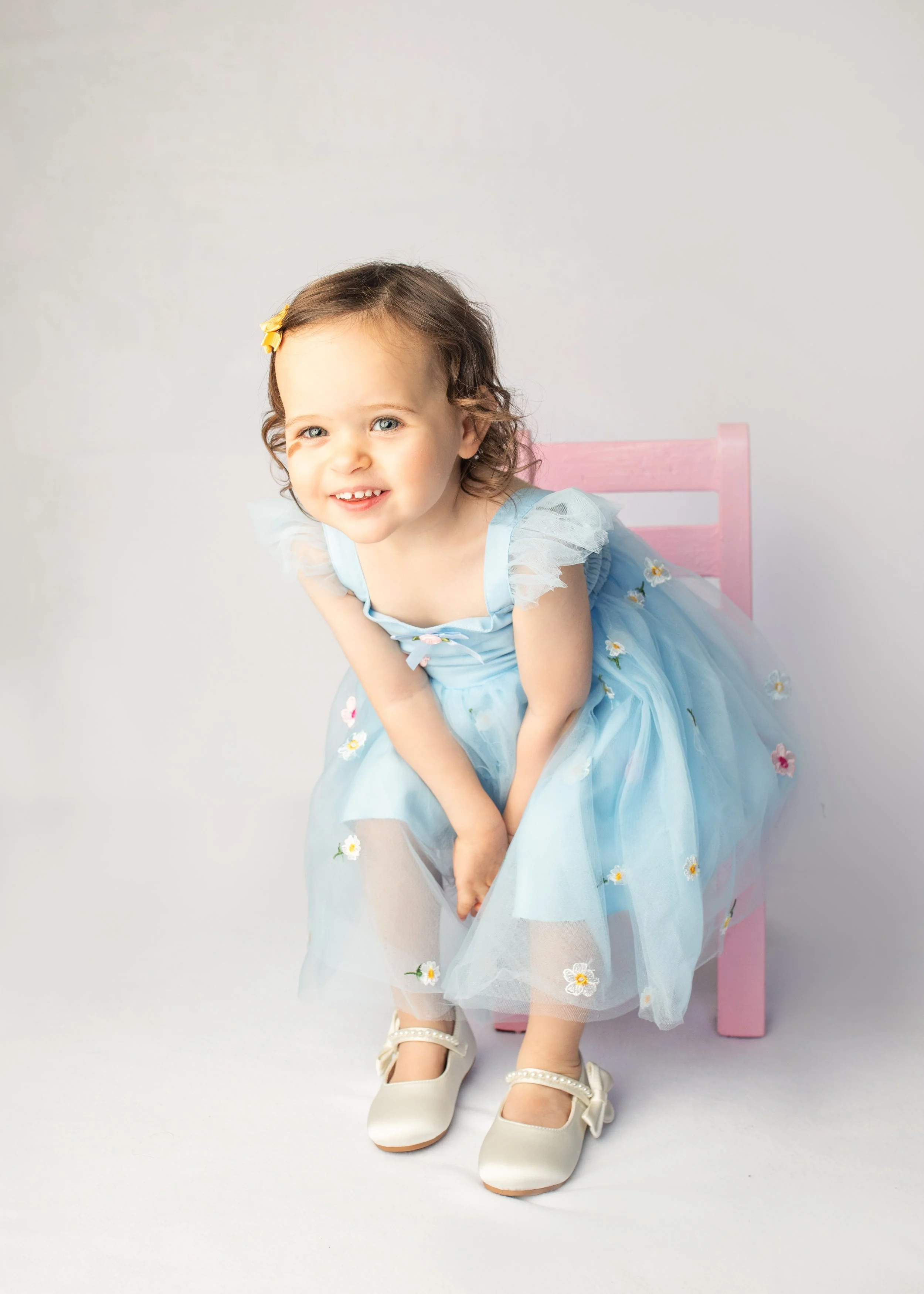 A young girl with light brown, curly hair sitting on a pink chair, wearing a light blue dress with floral embroidery, white shoes, and a yellow hair clip. She is smiling and leaning forward.